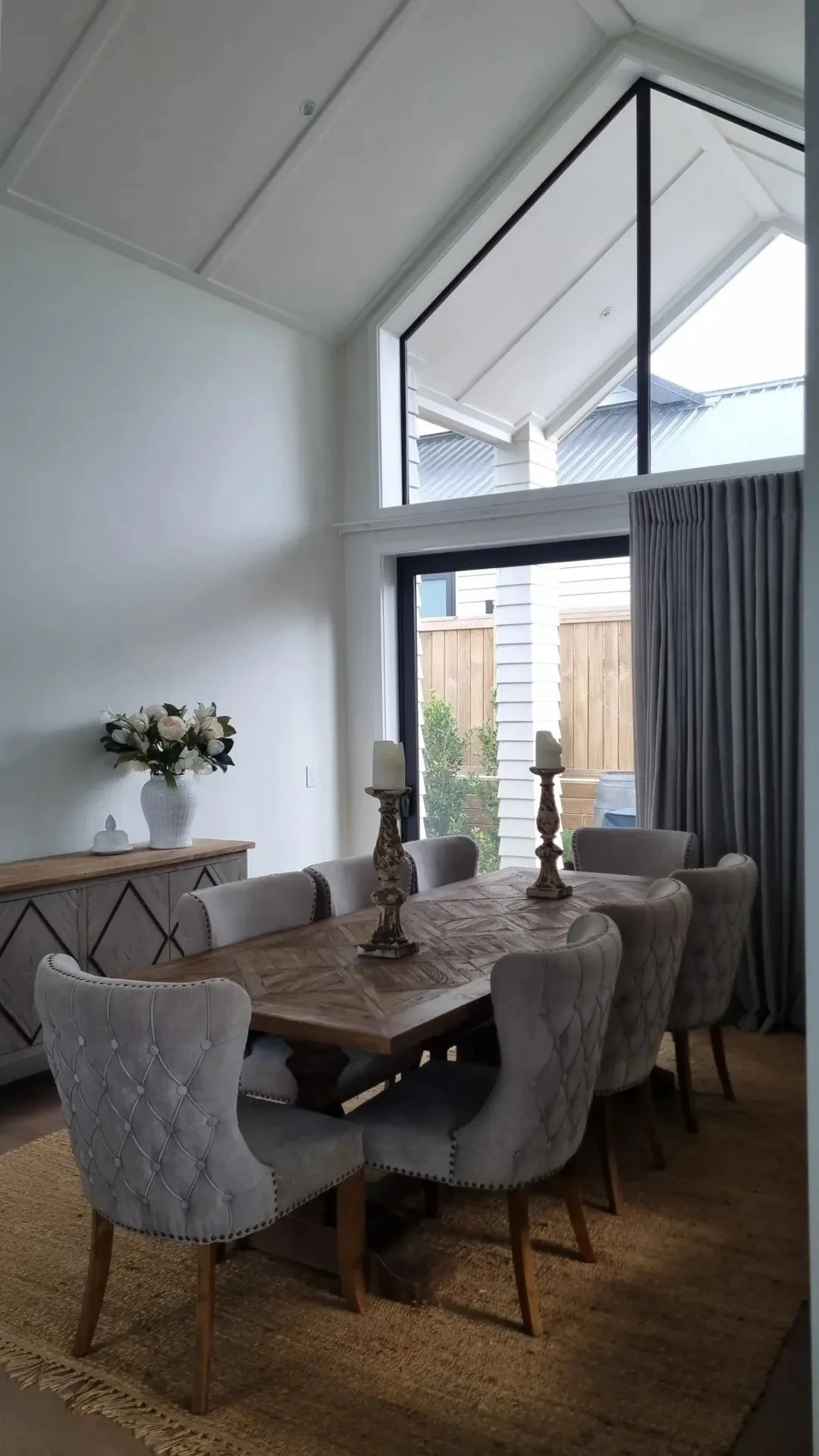 A dining room featuring a wooden dining table with six upholstered chairs, two candle holders with candles on the table, a sideboard with a vase of white roses, large windows with curtains, and a view of a backyard with a wooden fence and trees.