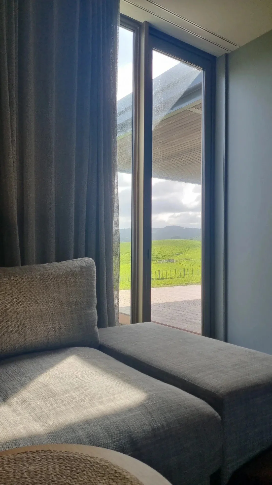 The photo shows a beige sofa in a room, with a glass sliding door opening to a scenic outdoor view of green rolling hills and a cloudy sky. Blue curtains are partially drawn to the side.