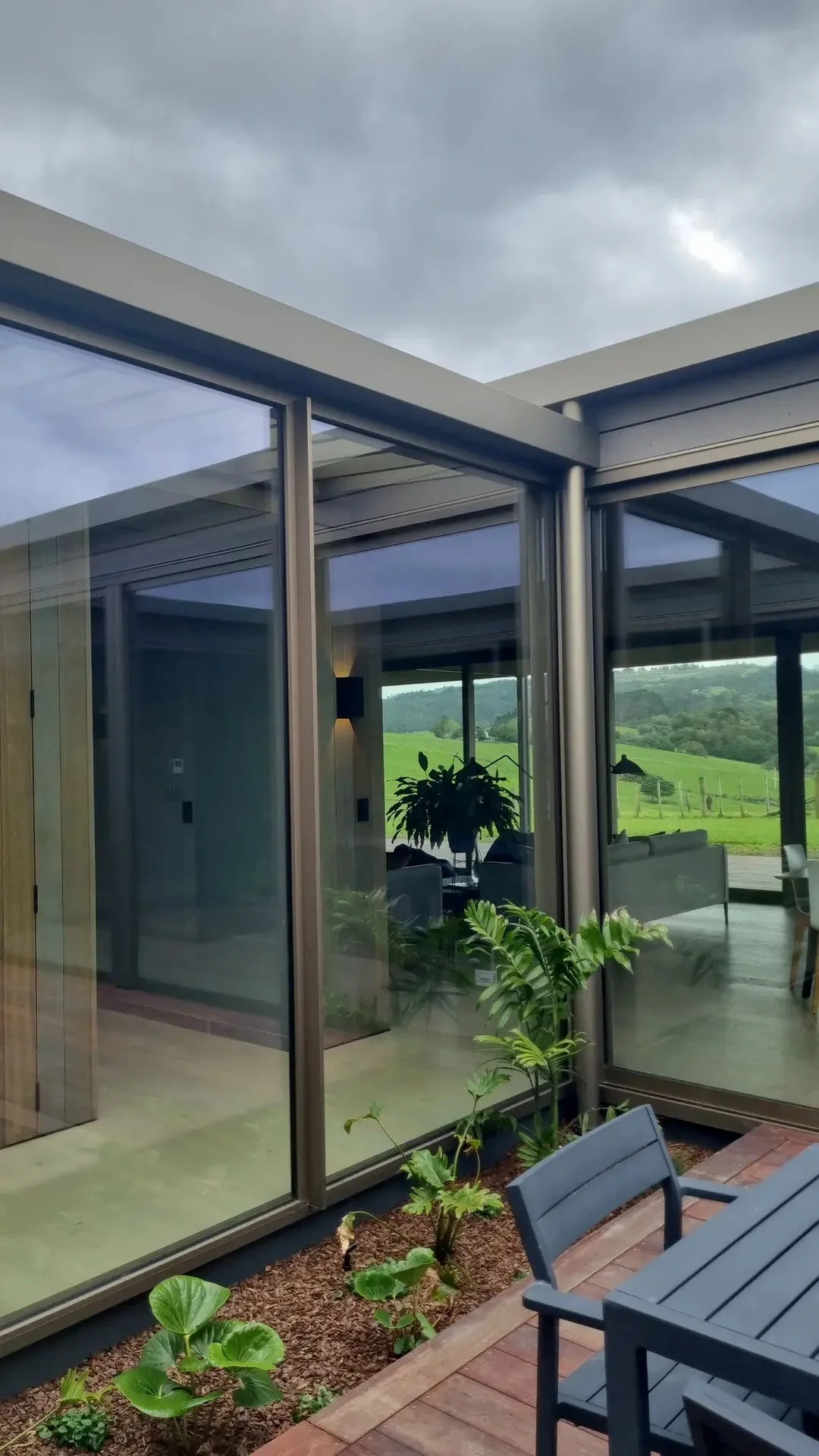 A modern house with large glass windows and doors, overlooking a lush green landscape with rolling hills and a cloudy sky.