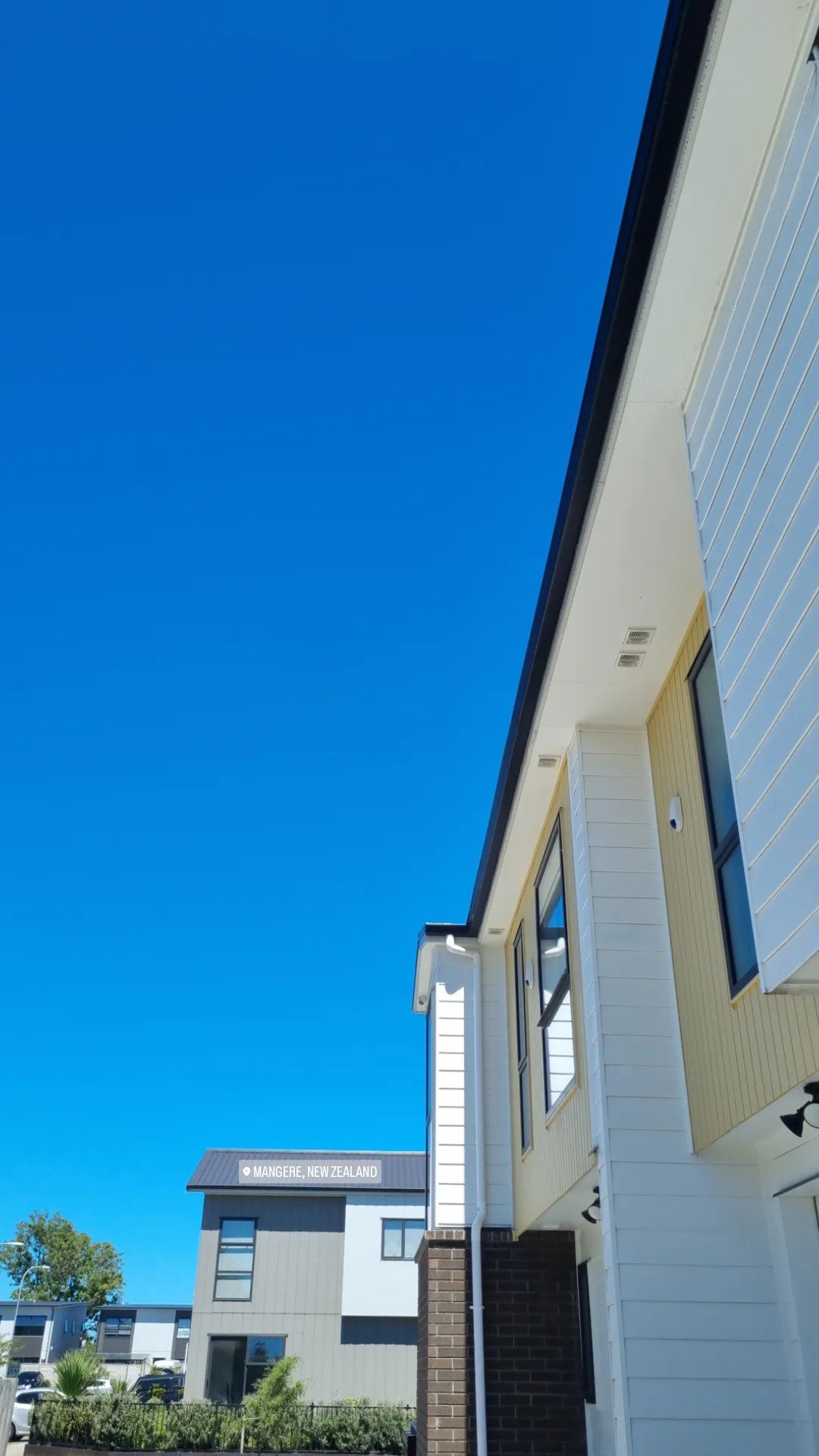 View of modern houses in Mangere, New Zealand under a clear blue sky, featuring the exterior of a yellow and white house with large windows.