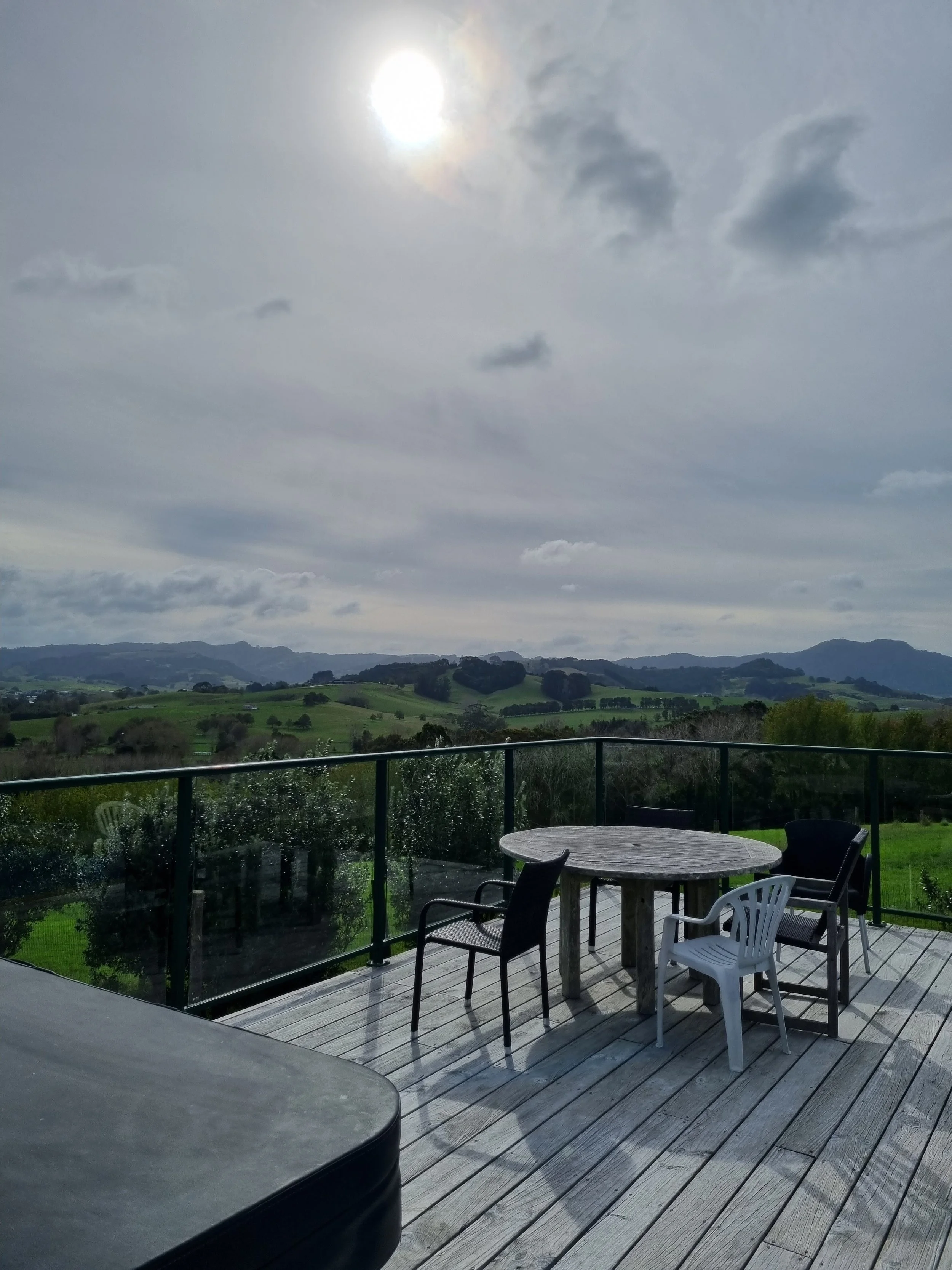 Outdoor terrace with a round wooden table and plastic chairs, overlooking rolling green hills under a cloudy sky with the sun partially obscured.