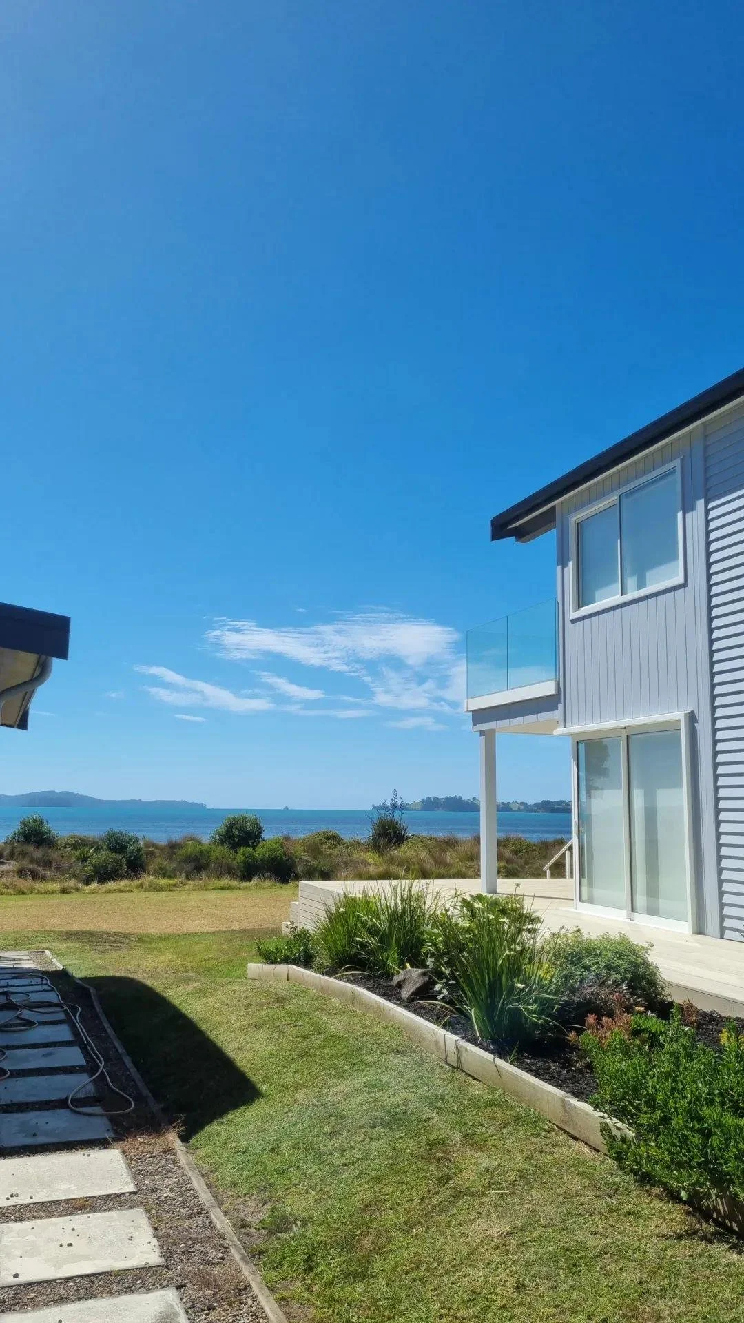 A modern house with a small balcony overlooking a grassy yard, with a view of the ocean and islands in the distance, under a clear blue sky.