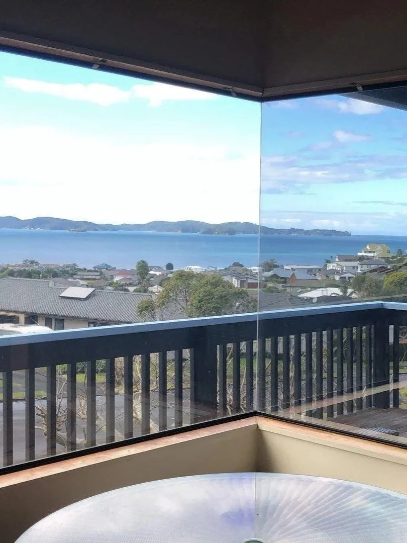 View of a town, water, and distant hills through a large glass window from a balcony.