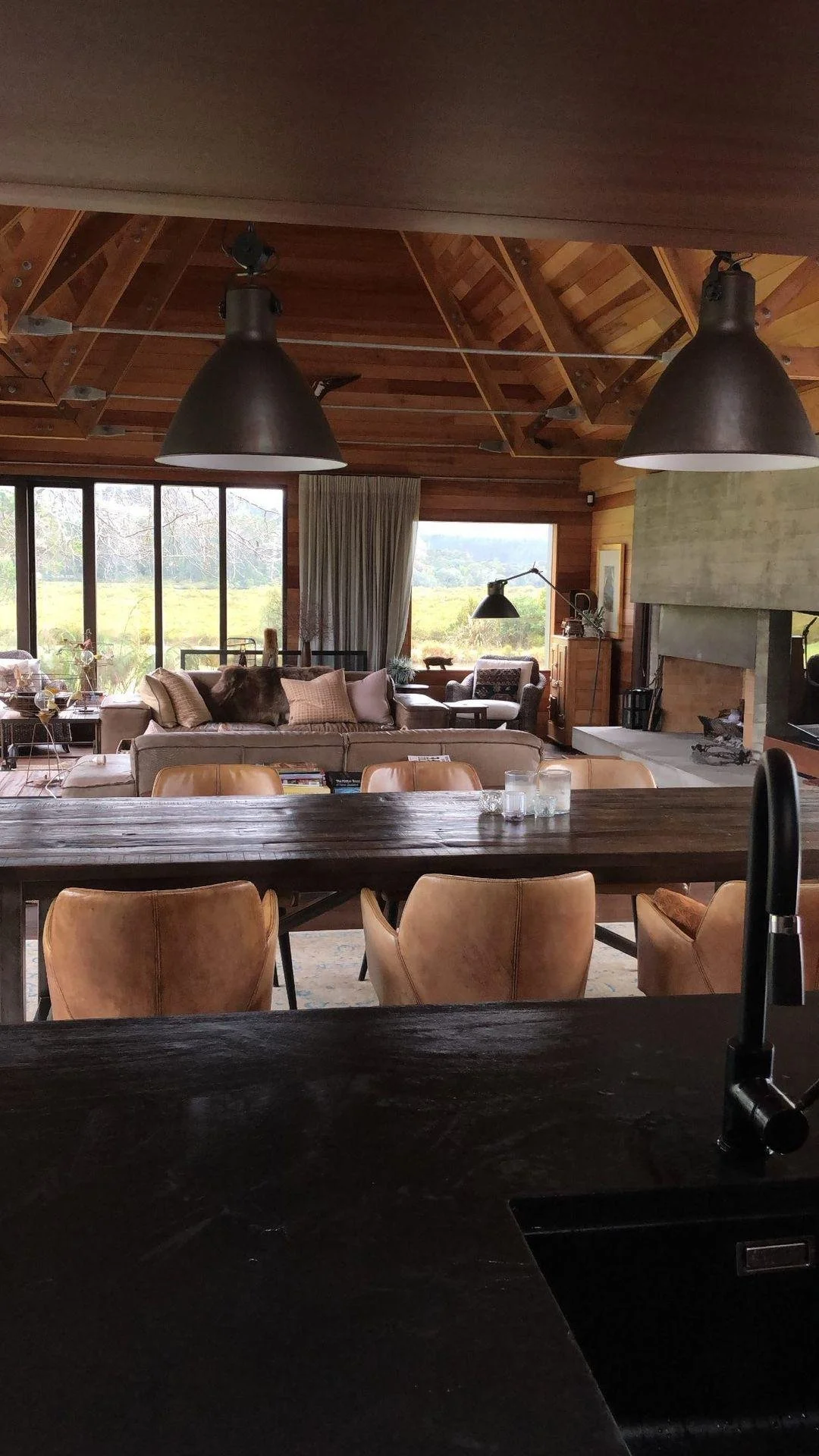 Living room with large windows, wooden ceiling, beige sofa, leather chairs, fireplace, and wooden dining table.