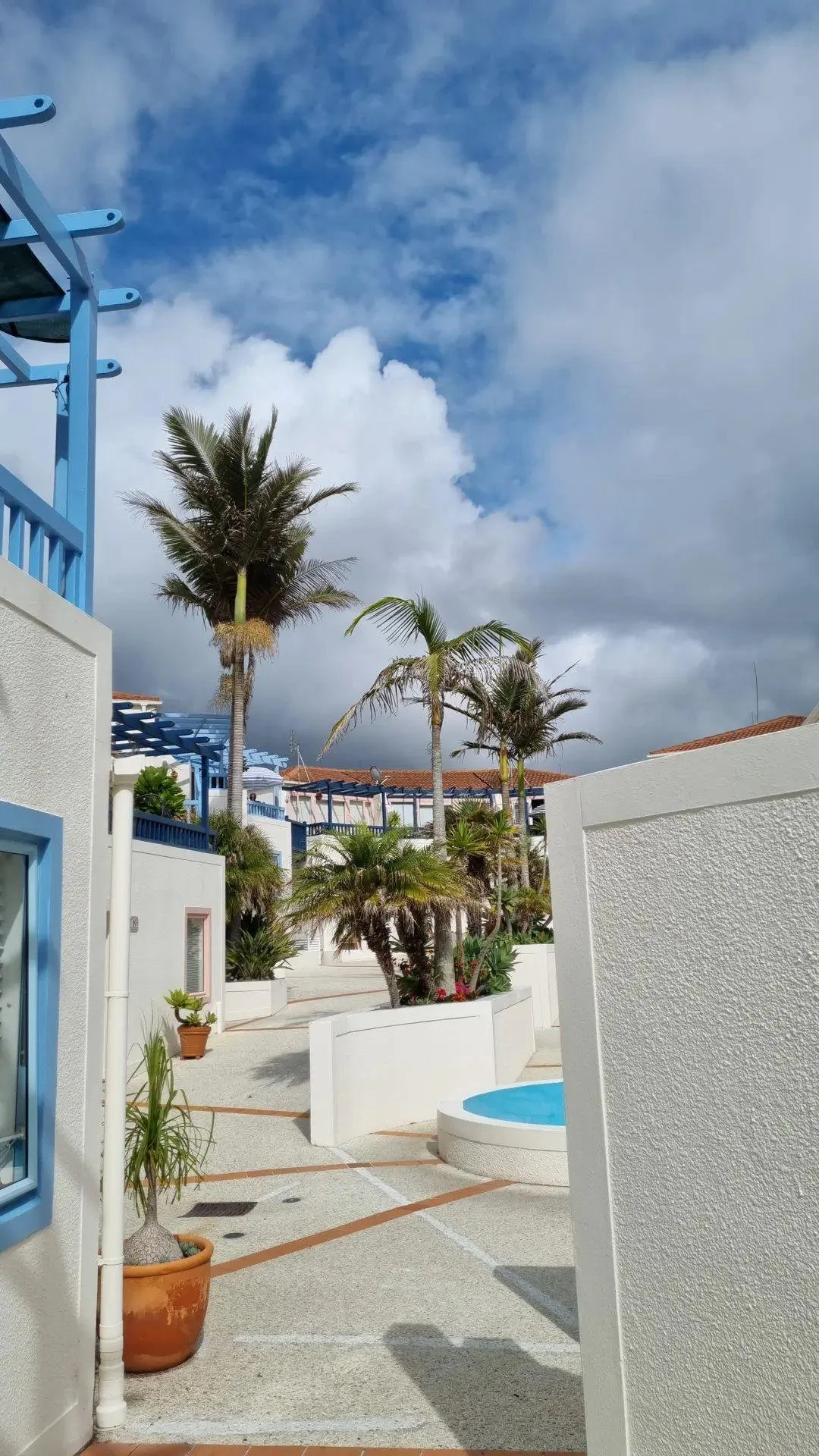 Tropical poolside area with white buildings, palm trees, and a partly cloudy sky.