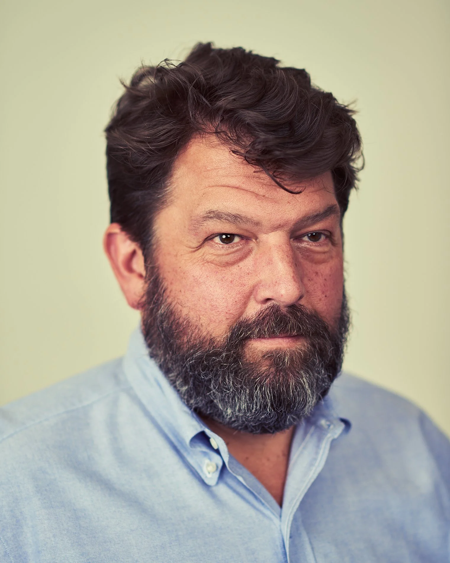A middle-aged man with dark, wavy hair and a full beard, wearing a light blue dress shirt, looking at the camera against a plain, light-colored background.