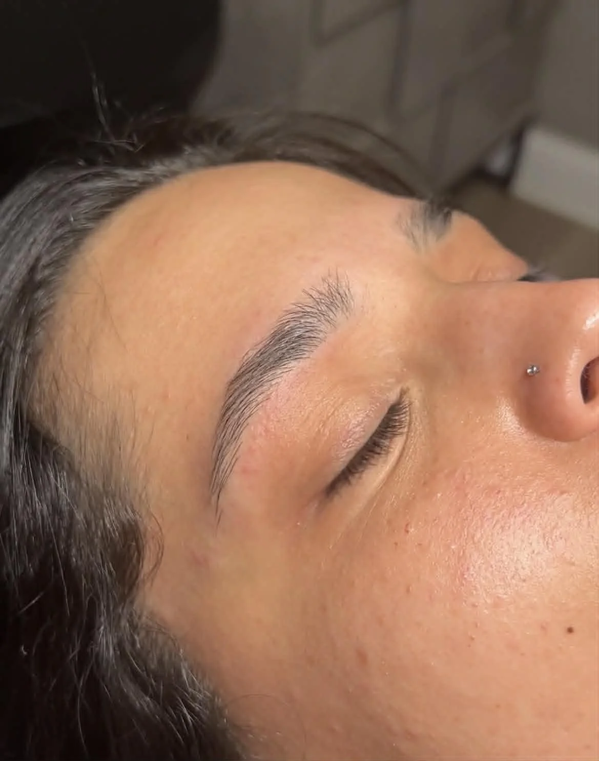 Close-up of a woman lying down with eyes closed, showing well-groomed eyebrows and a nose piercing.