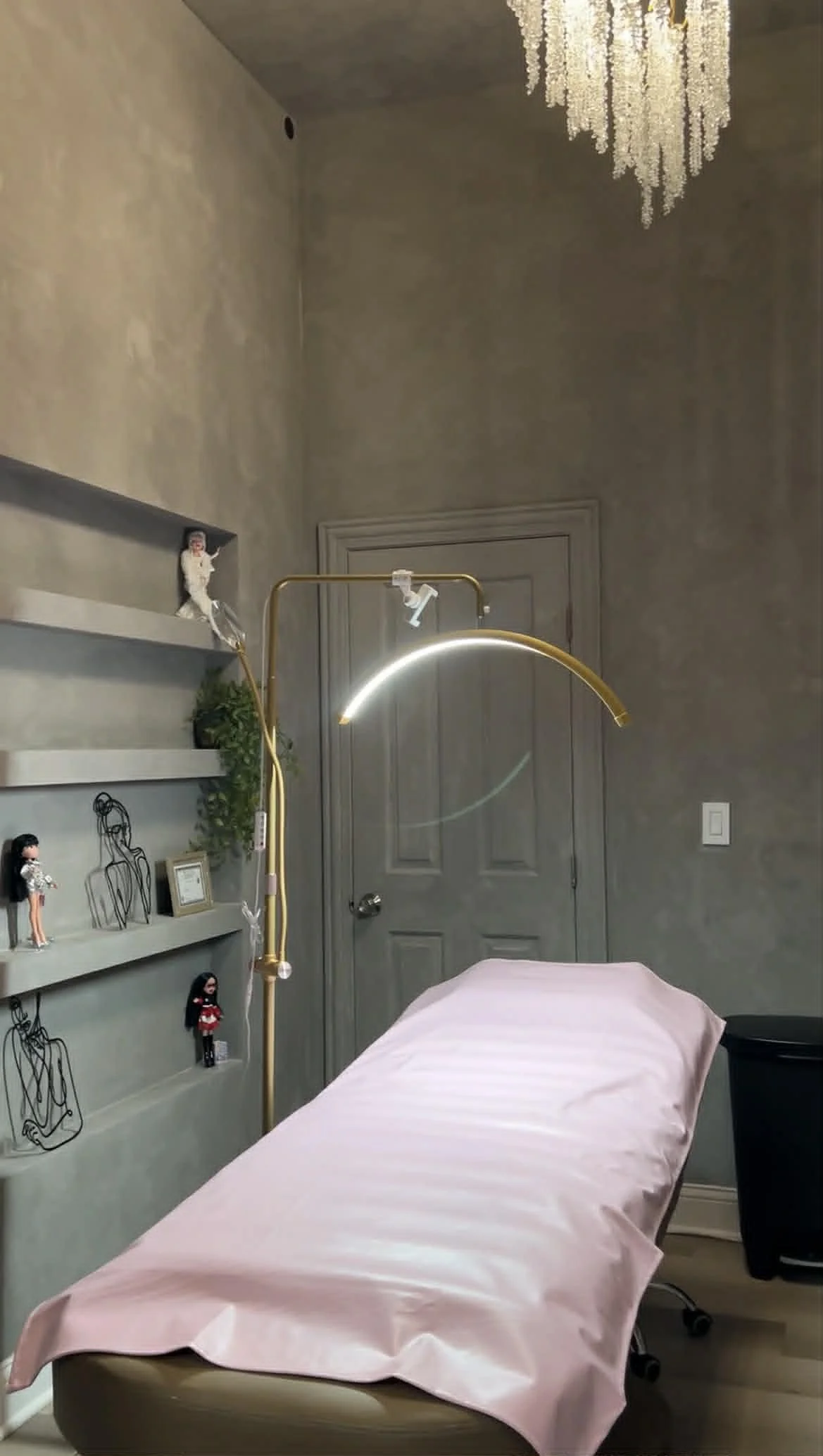 A medical or spa examination room with a pink draped table in the foreground. A gold-colored adjustable lamp is positioned over the table. On the left wall, there are shelves with various figurines, wire art, and framed photos. The room has beige wal
