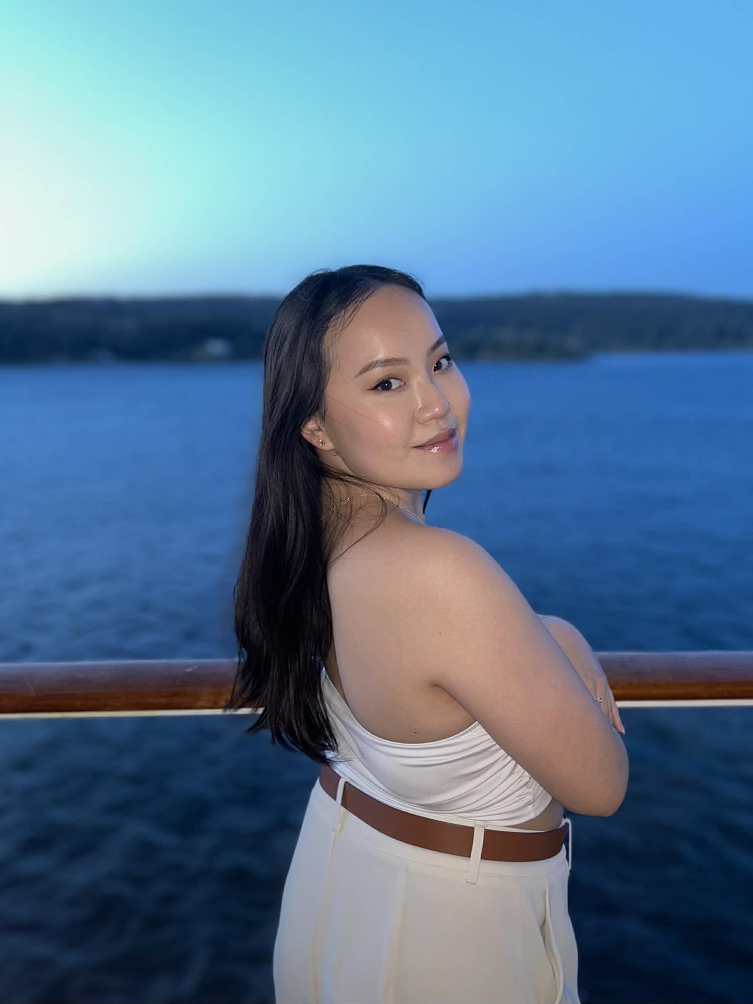 A woman with long dark hair and a light complexion posing with her back and shoulders exposed on a boat with a blue body of water and distant land in the background.