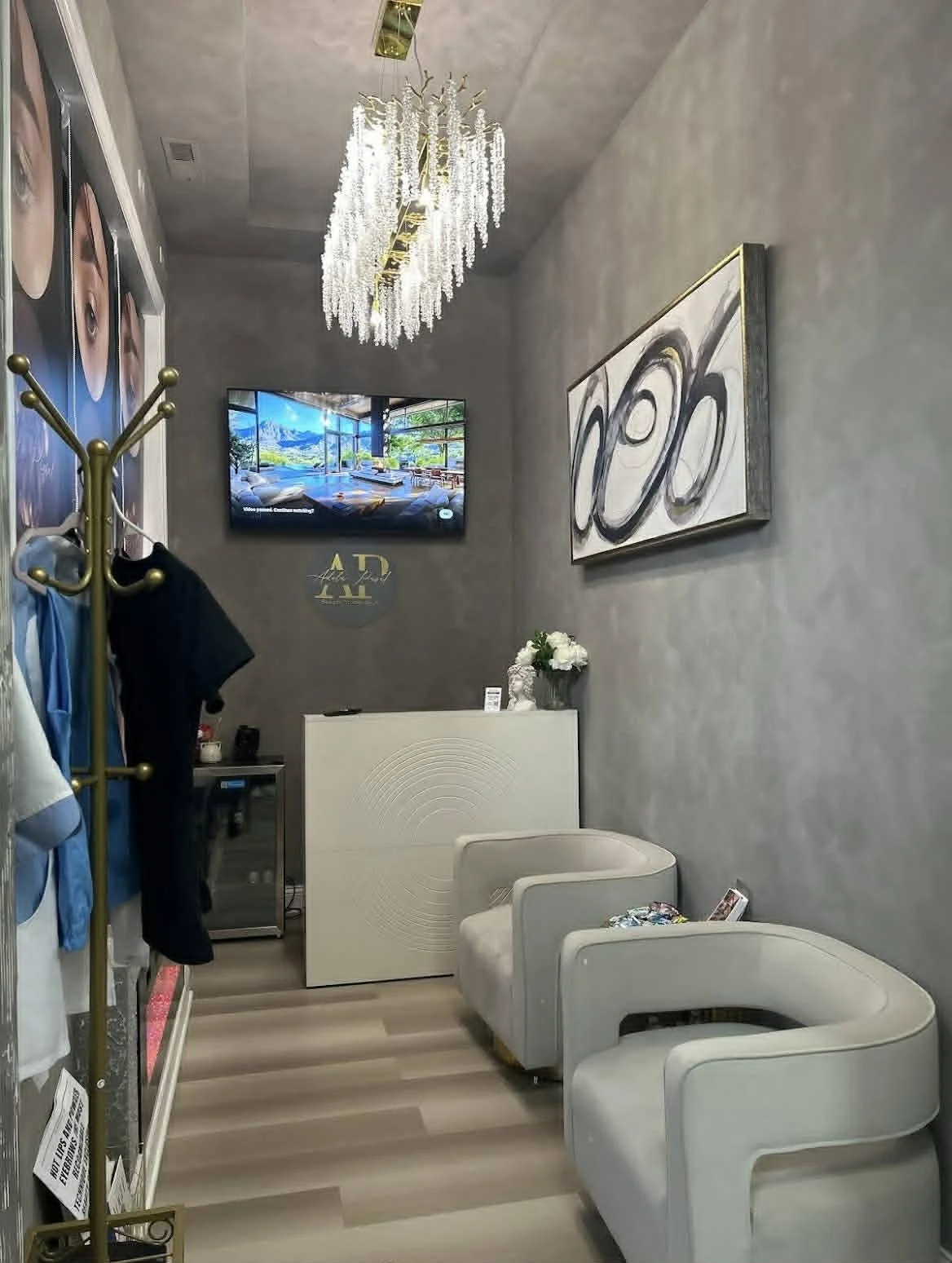 Interior of a waiting area with two cream-colored armchairs, a white reception desk with a flower vase, abstract black and white artwork on gray walls, a chandelier hanging from the ceiling, a wall-mounted TV showing a scenic landscape, and a coat ra