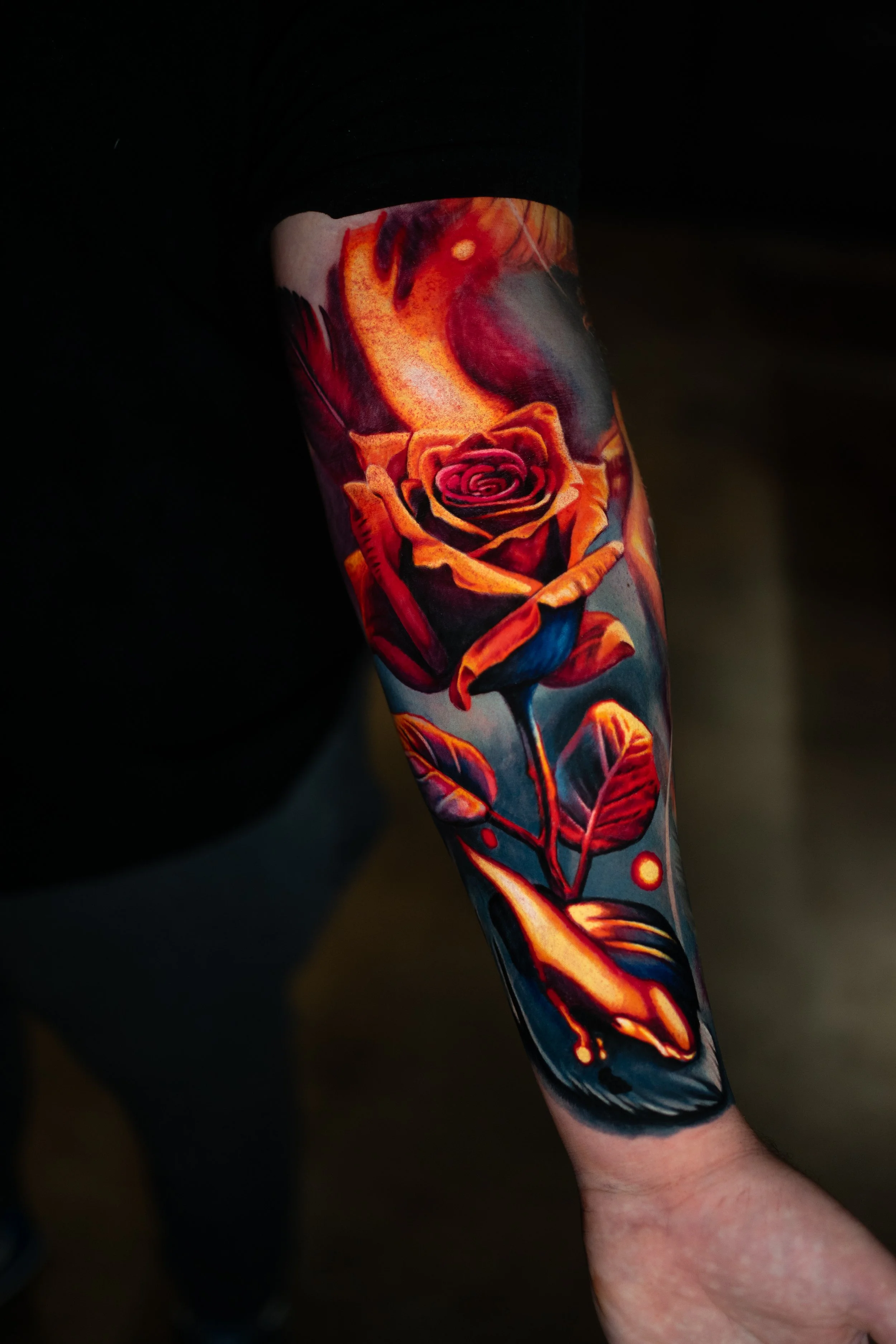 Ein Arm mit einem detaillierten, bunten Tattoo eines roten Rosenstrauchs mit Blättern. Im Hintergrund ist die Hand des Tattoo-Künstlers sichtbar.