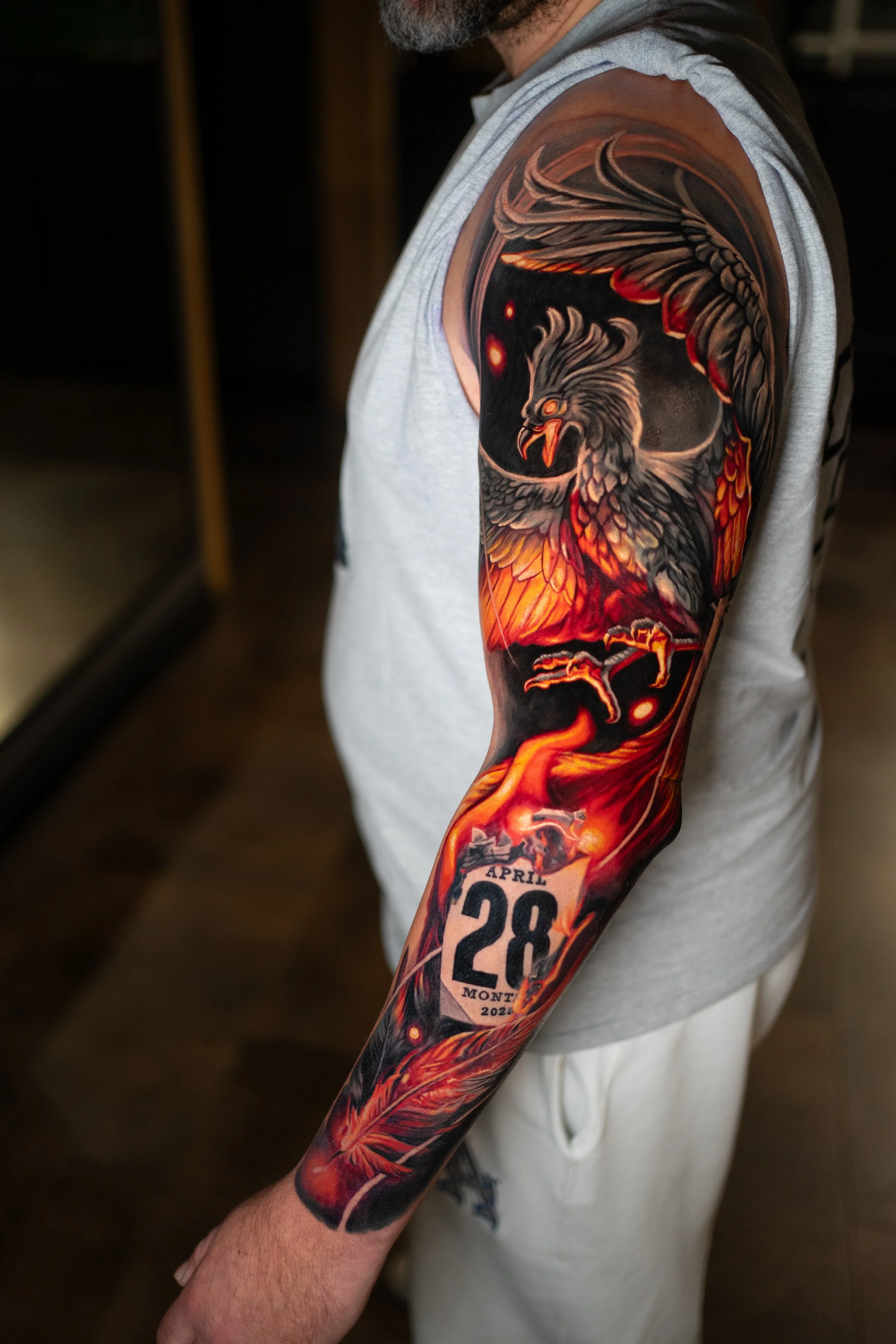Detailliertes Tattoo eines fliegenden Adlers mit Feuer- und Galaxie-Motiven auf dem Arm eines Mannes, inklusive Datum und Ort. Der Mann trägt ein graues T-Shirt.