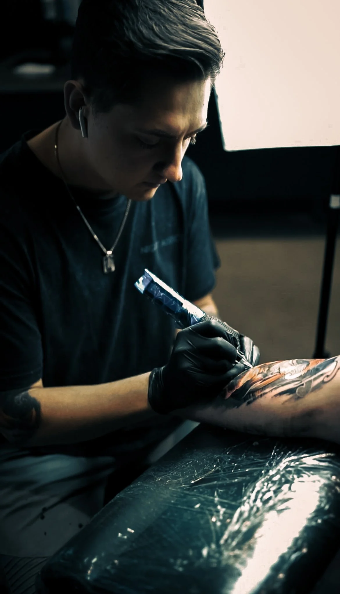 Ein junger Mann tätowiert mit schwarzen Handschuhen ein Tattoo auf dem Unterarm. Er trägt ein schwarzes T-Shirt, eine Halskette, und hat kurze, helle Haare.