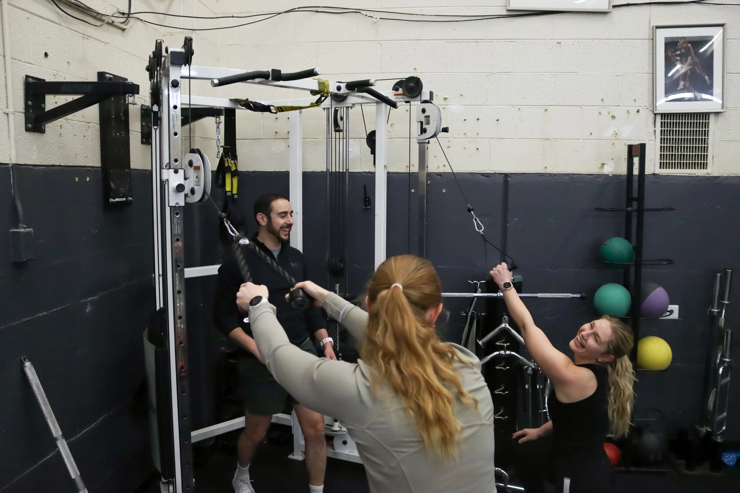 partner training session with two clients performing different exercises using cable machines while coach guides them.