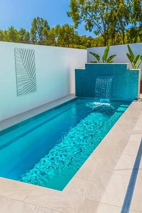 A modern backyard pool with a water feature and white walls, surrounded by green trees and plants.