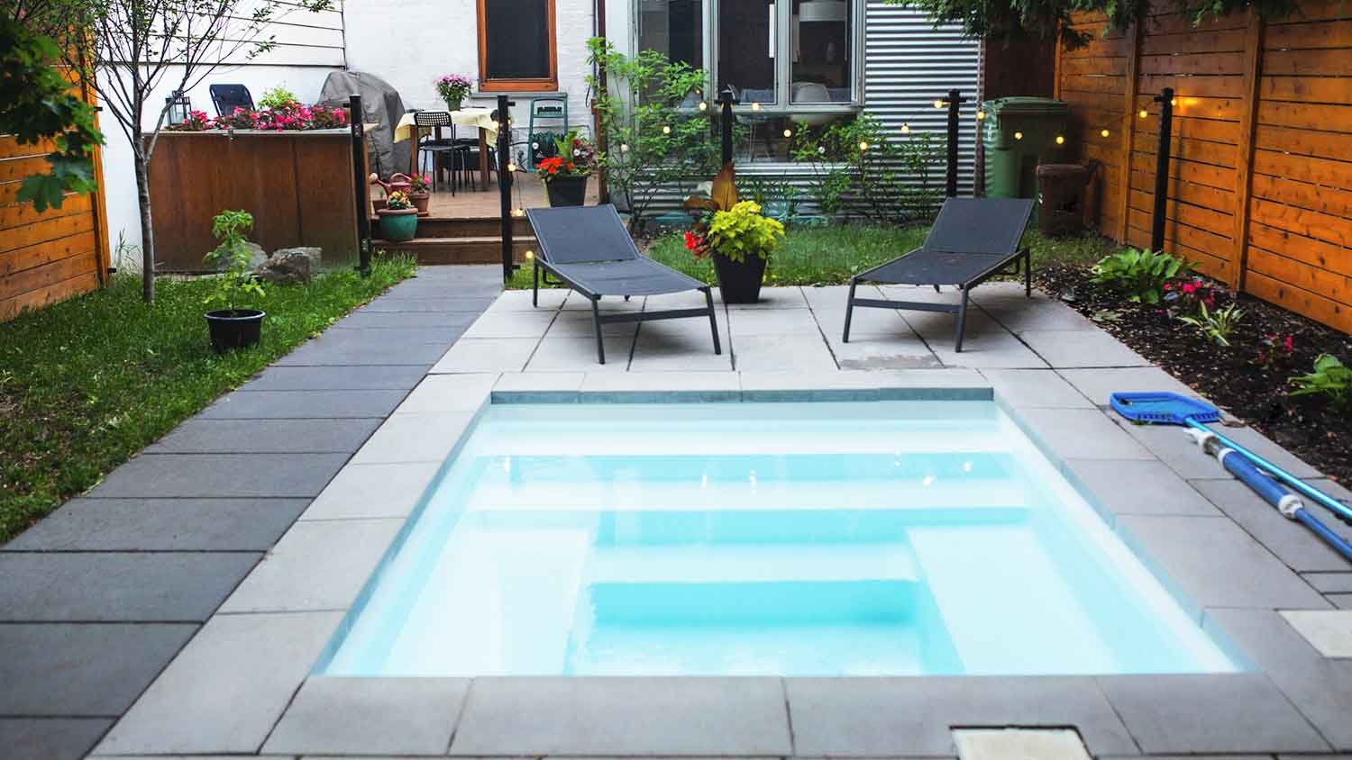 A backyard with a small illuminated pool, two lounge chairs, potted plants, a flower bed, and a privacy fence.