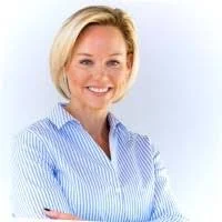 Portrait of a woman with blonde hair wearing a light blue striped shirt, smiling with arms crossed.