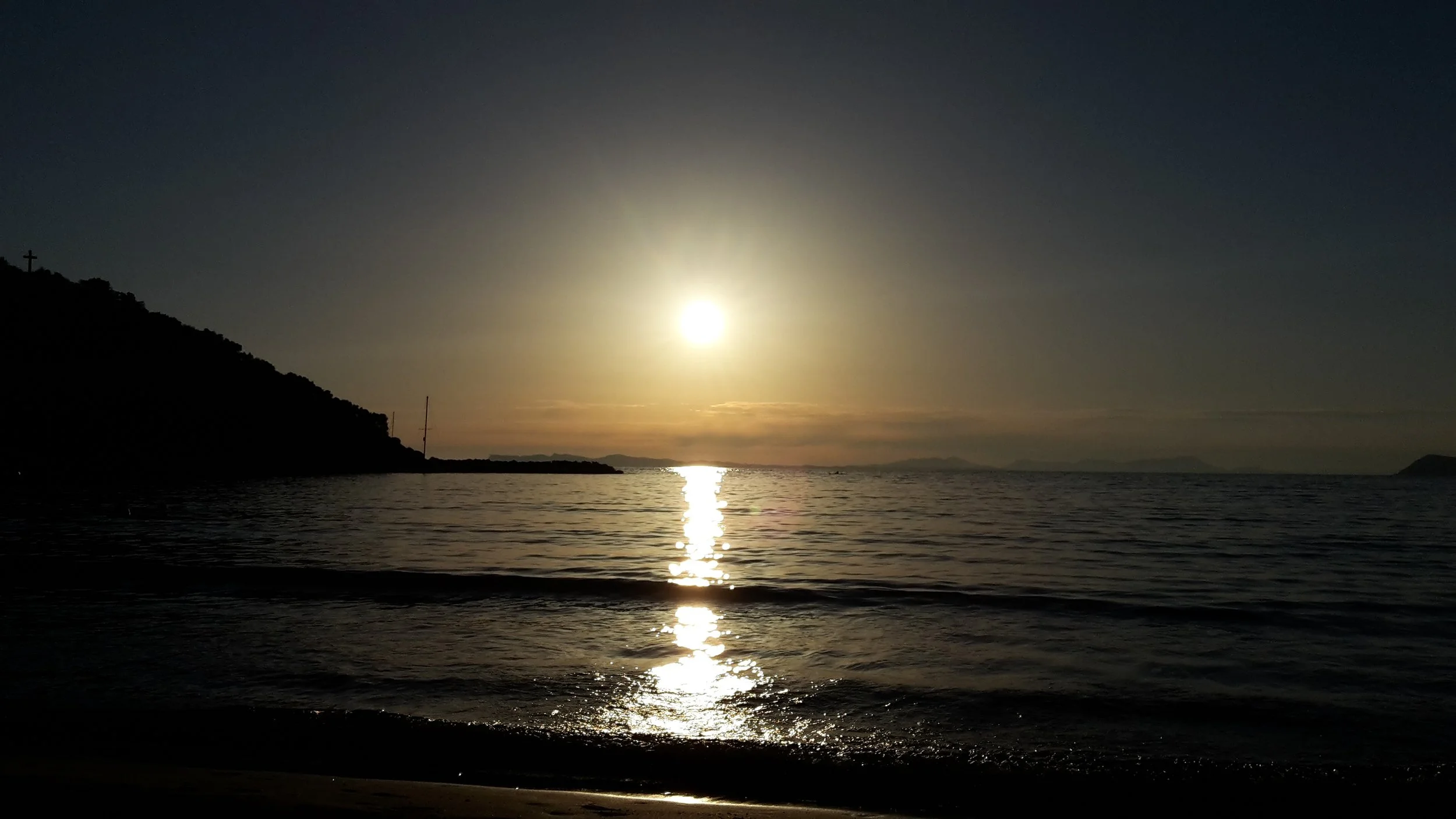 A scenic shot of a bright, brilliant sun high above the ocean, casting a sharp, sparkling silver reflection on the dark water's surface.