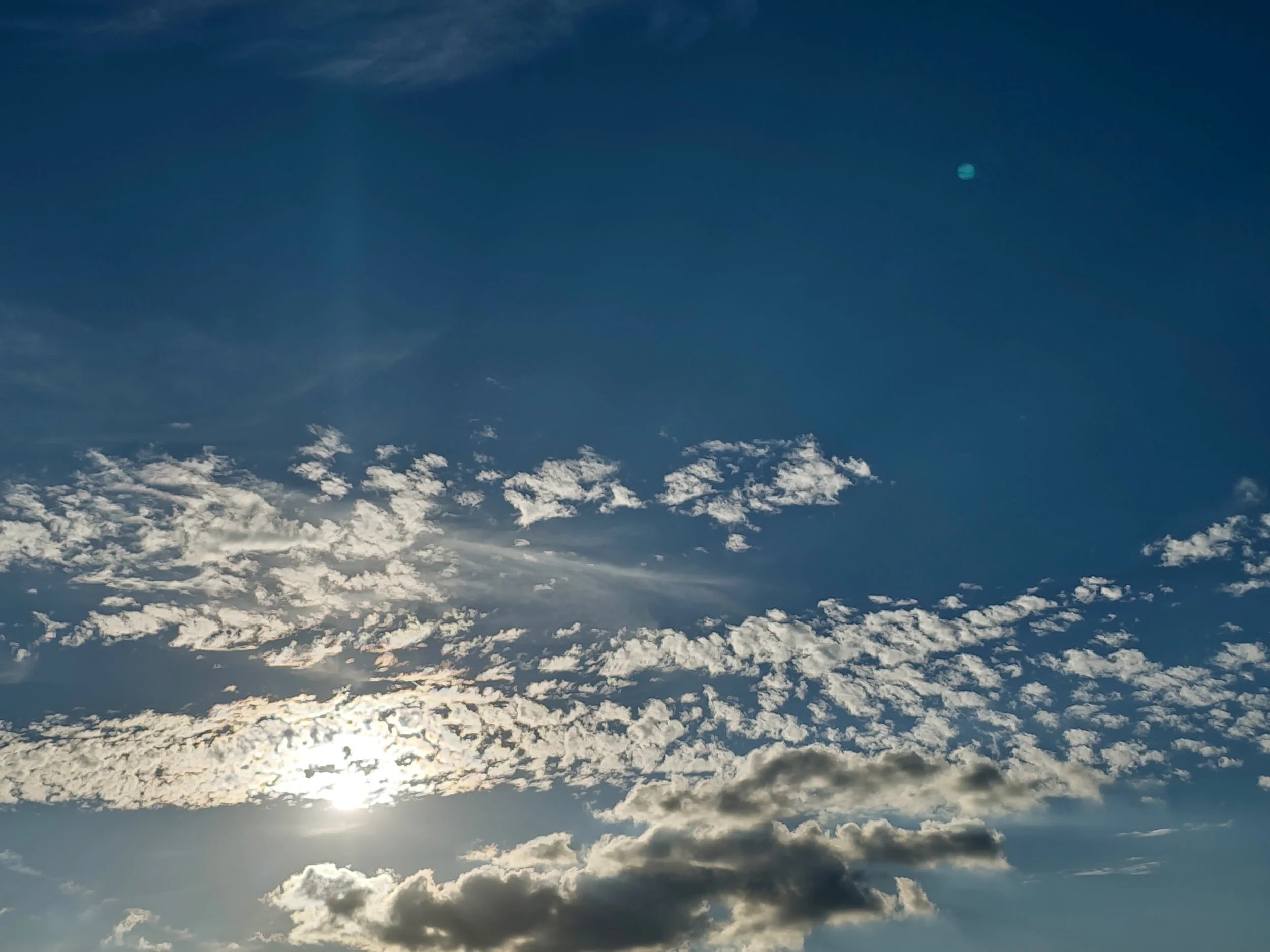 The bright sun shining through thin, scattered white clouds across a clear blue afternoon sky.