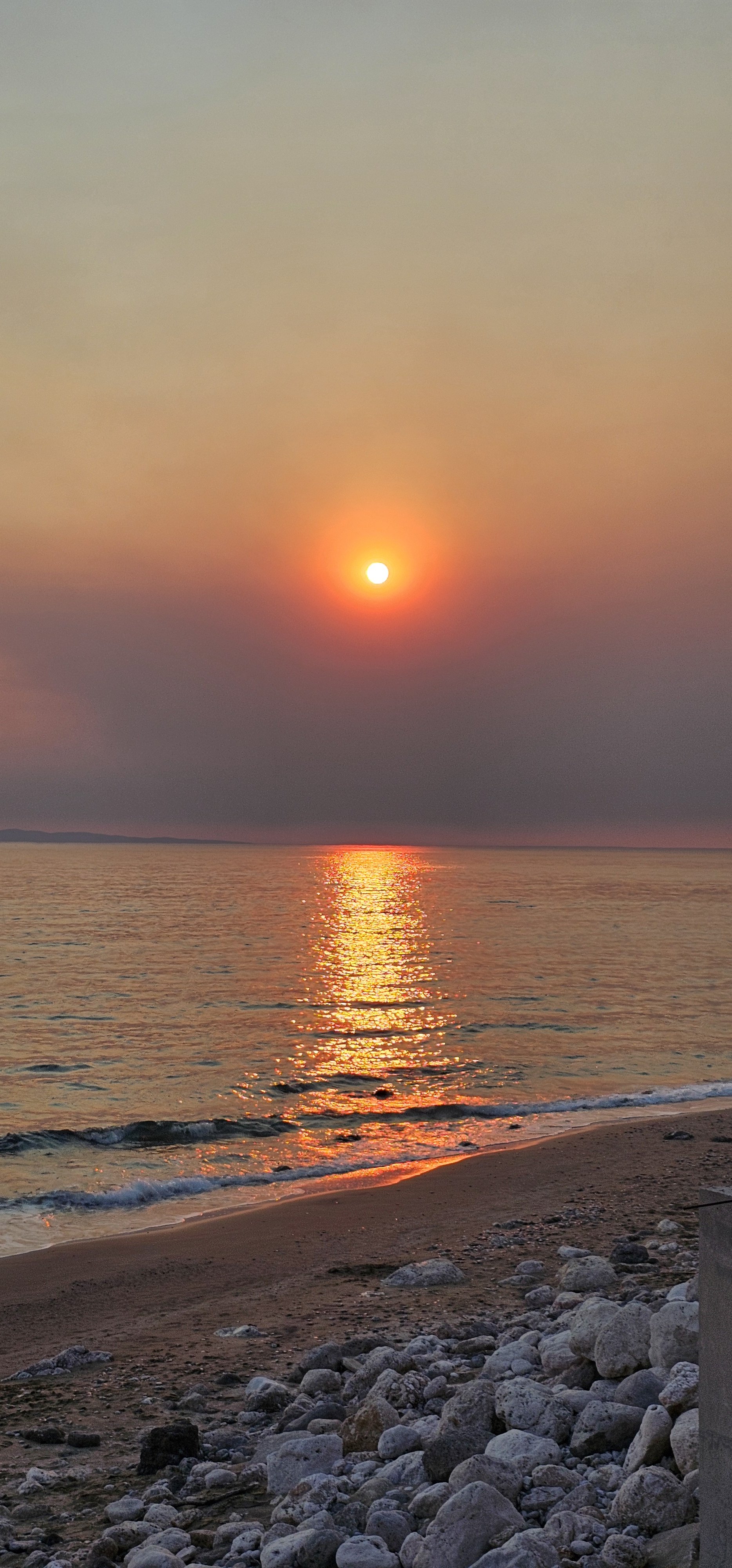 A vertical photograph of a glowing orange sun setting over the ocean, casting a vibrant golden reflection across the water onto a rocky beach.