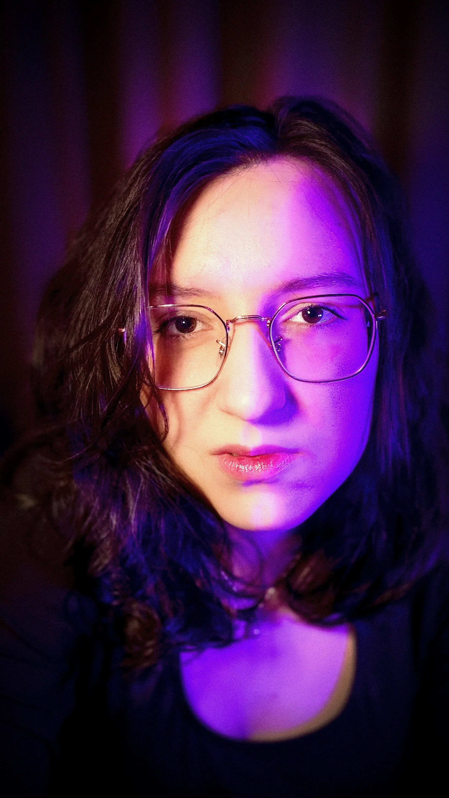 A close-up portrait of singer, songwriter, and producer Sandra Spirou. She has dark wavy hair, wears wire-rimmed glasses, and is dramatically lit by intense purple and pink neon light against a dark background.
