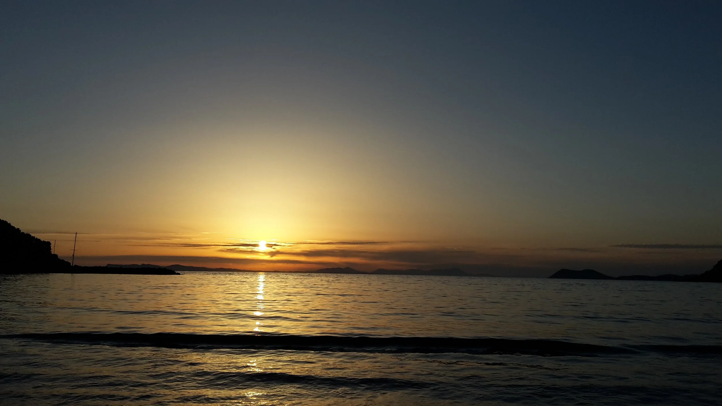A wide landscape of a bright sun setting low over the calm sea, creating a golden horizon between dark silhouettes of distant land.