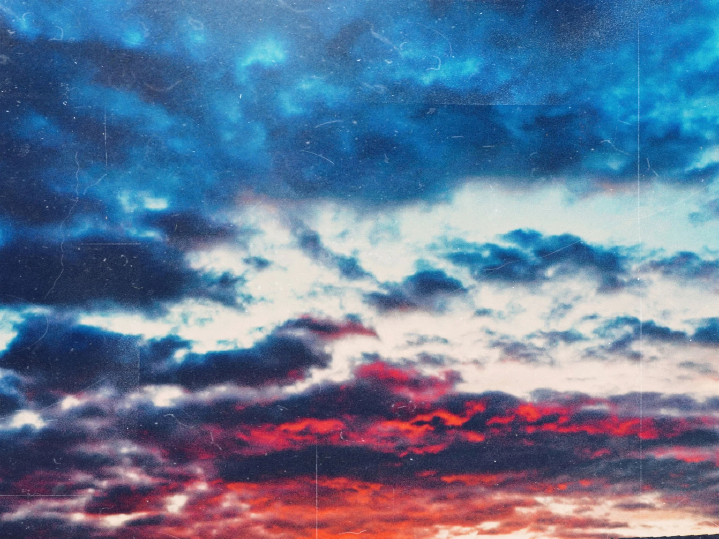 A dramatic sunset photograph showing thick, dark clouds heavily layered with deep blue tones, contrasting sharply with fiery red and orange colors along the lower horizon.