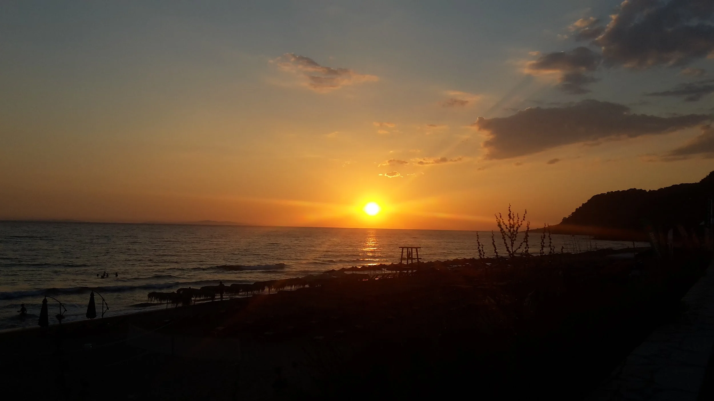 A coastal sunset showing a bright sun dipping towards the horizon over the sea, with dark silhouettes of a beach and a hillside on the right.