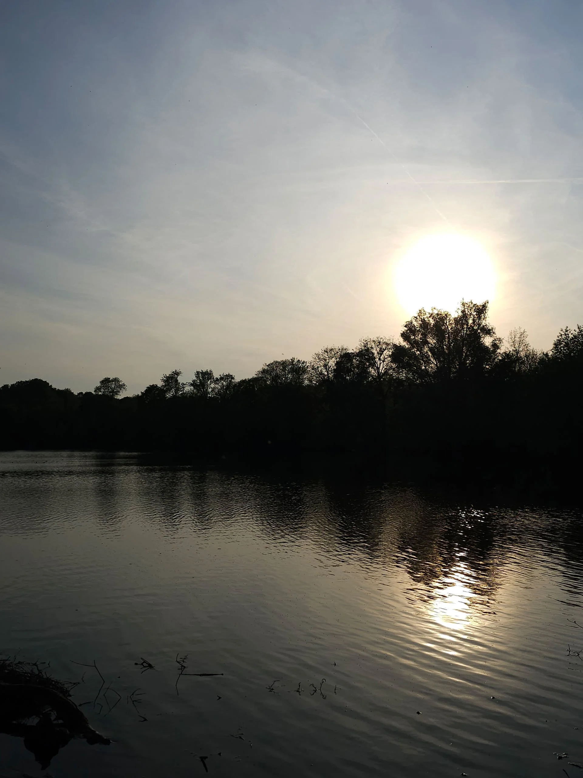 A scenic photograph of a bright sun setting over a quiet lake, creating a vibrant golden reflection on the water's surface beneath a silhouette of distant trees.