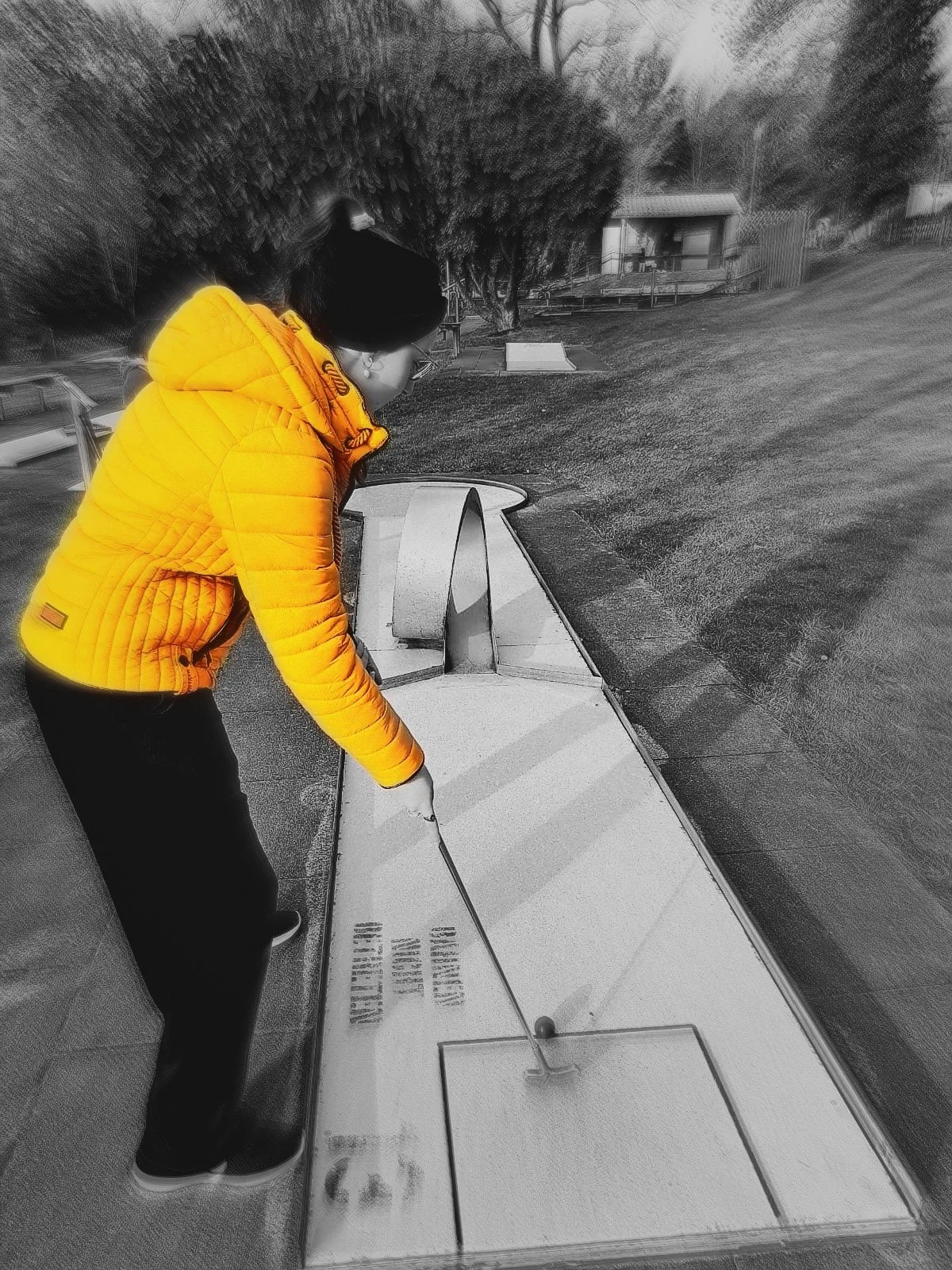 A stylized black and white photograph with a selective color effect, focusing on a person in a bright yellow jacket playing minigolf, enhanced by a dynamic motion blur around the edges.