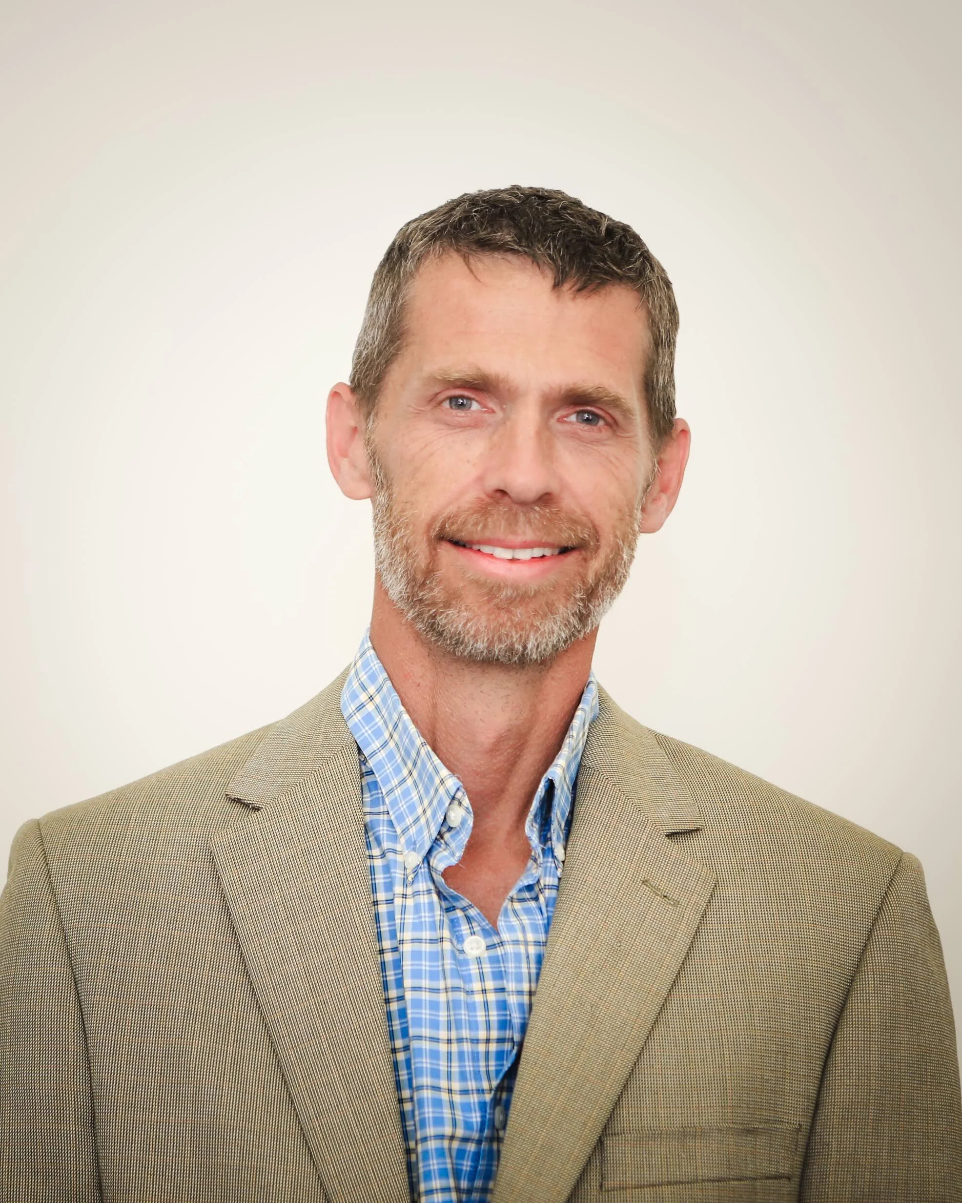 Close-up portrait of a smiling middle-aged man with short brown hair, blue eyes, and a gray beard, wearing a tan blazer and blue checkered shirt, against a plain white background.