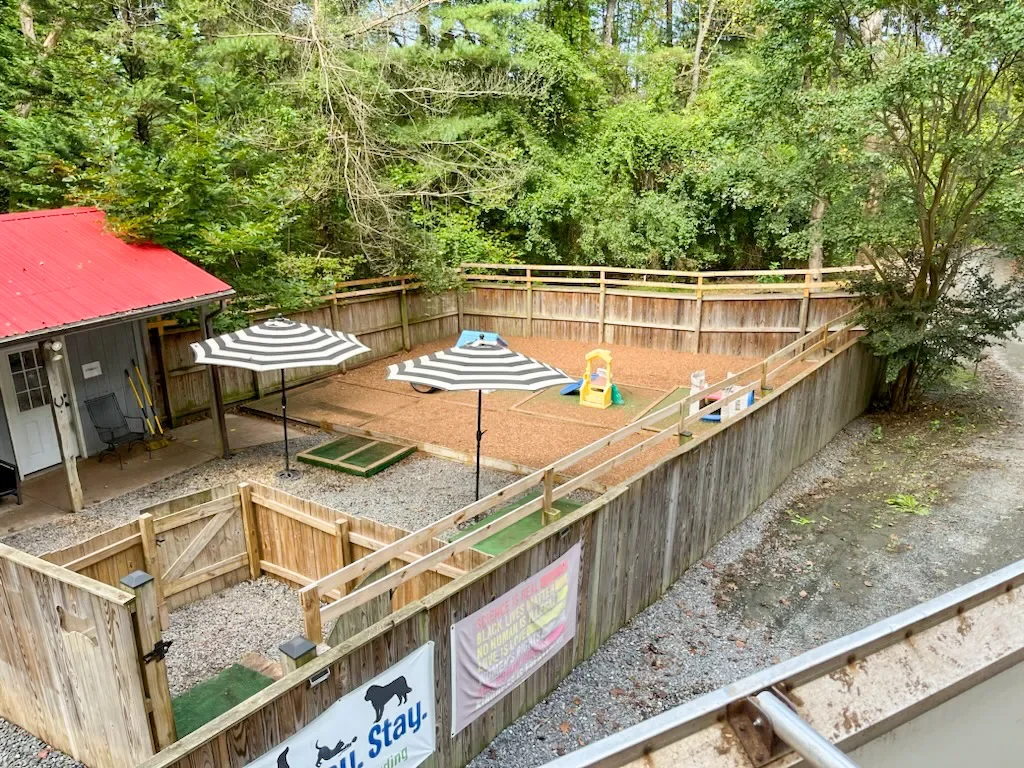 A fenced backyard with a play area that has a small slide and a sandbox, shaded by two umbrellas, neighboring a building with a red roof, with trees in the background.