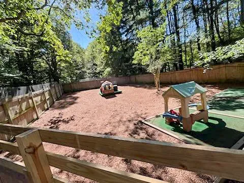 Large backyard with a fenced-in play area, sandbox, and small toy house, surrounded by trees.