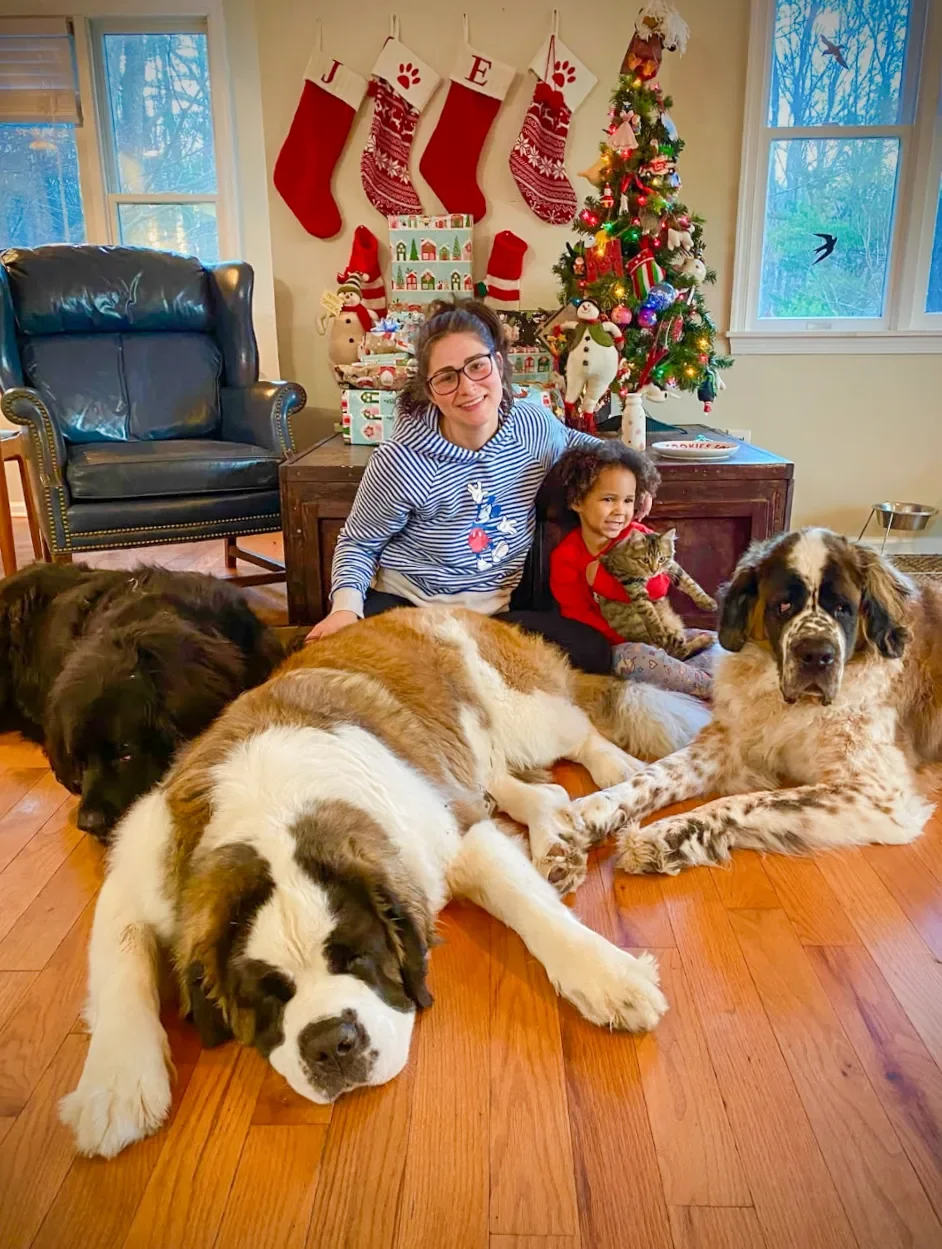 A woman with dark hair and glasses and a young girl sitting on the floor with three dogs and a cat in a festive living room decorated for Christmas, with a decorated tree, stockings, and wrapped presents.