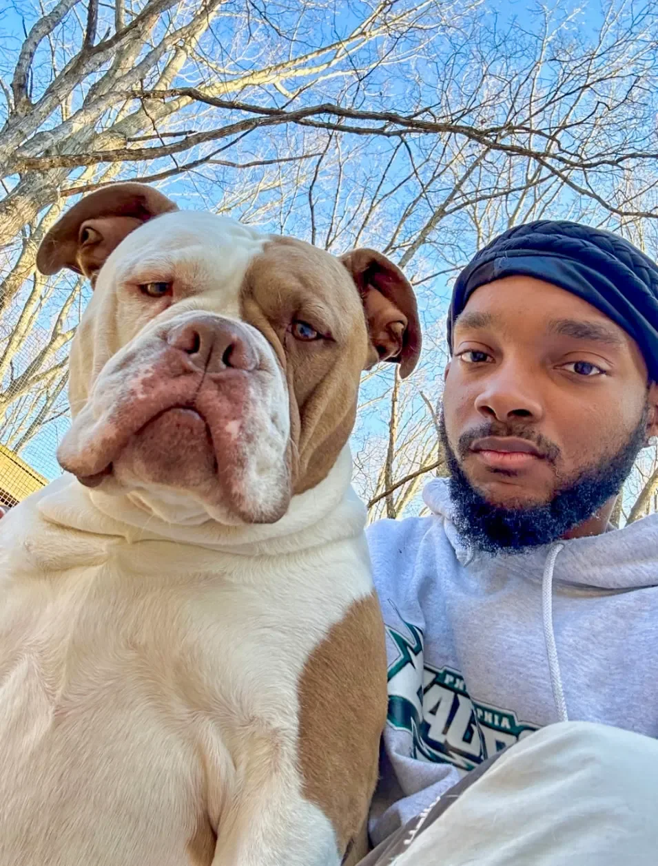 A young man with a beard and a dark blue head wrap  outdoors with a large bulldog resting against him. The background shows leafless trees and a clear blue sky.