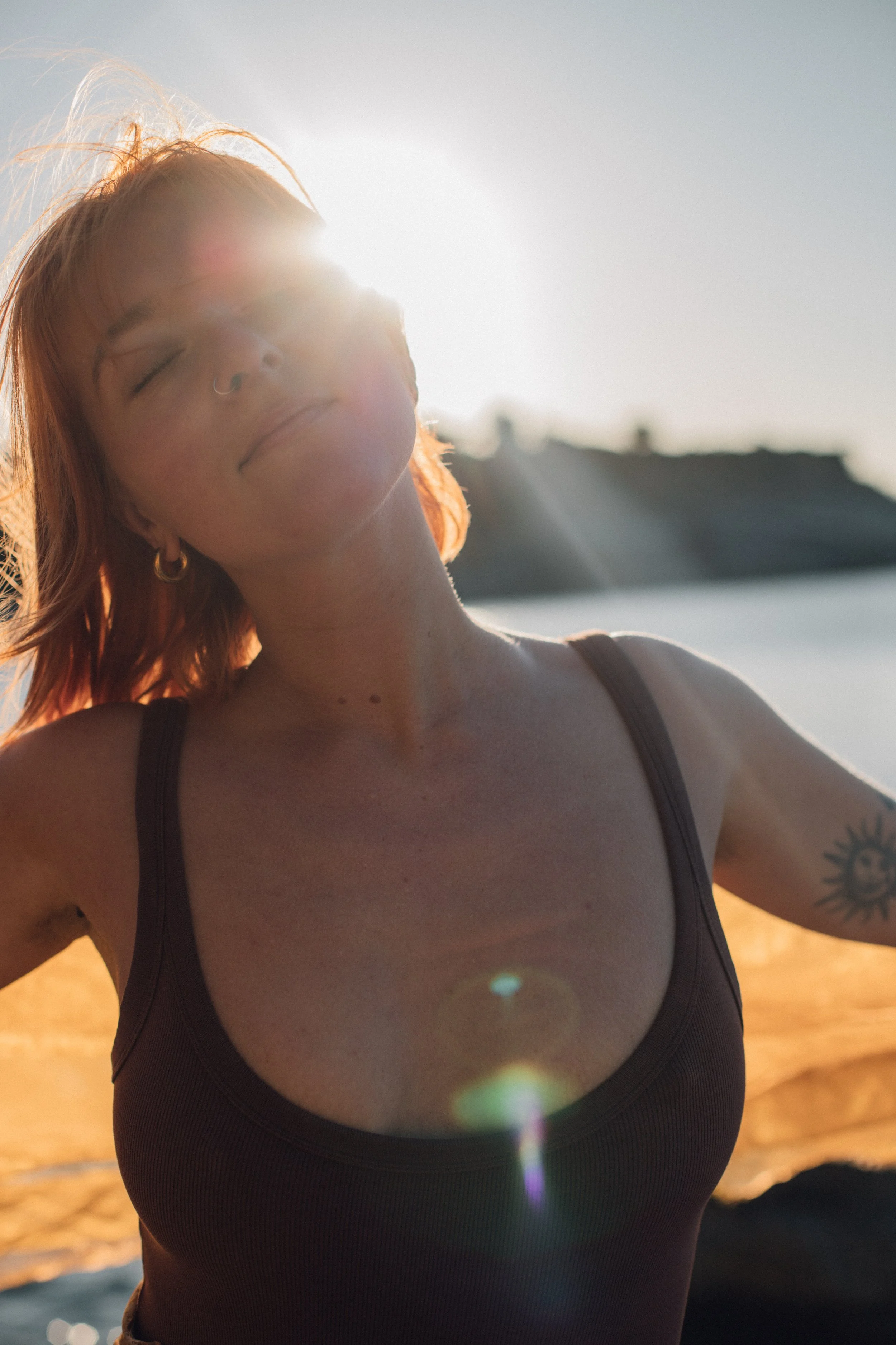 A woman with red hair, eyes closed, smiling, wearing a black top, standing outdoors with sunlight behind her, near water with rocks in the background.