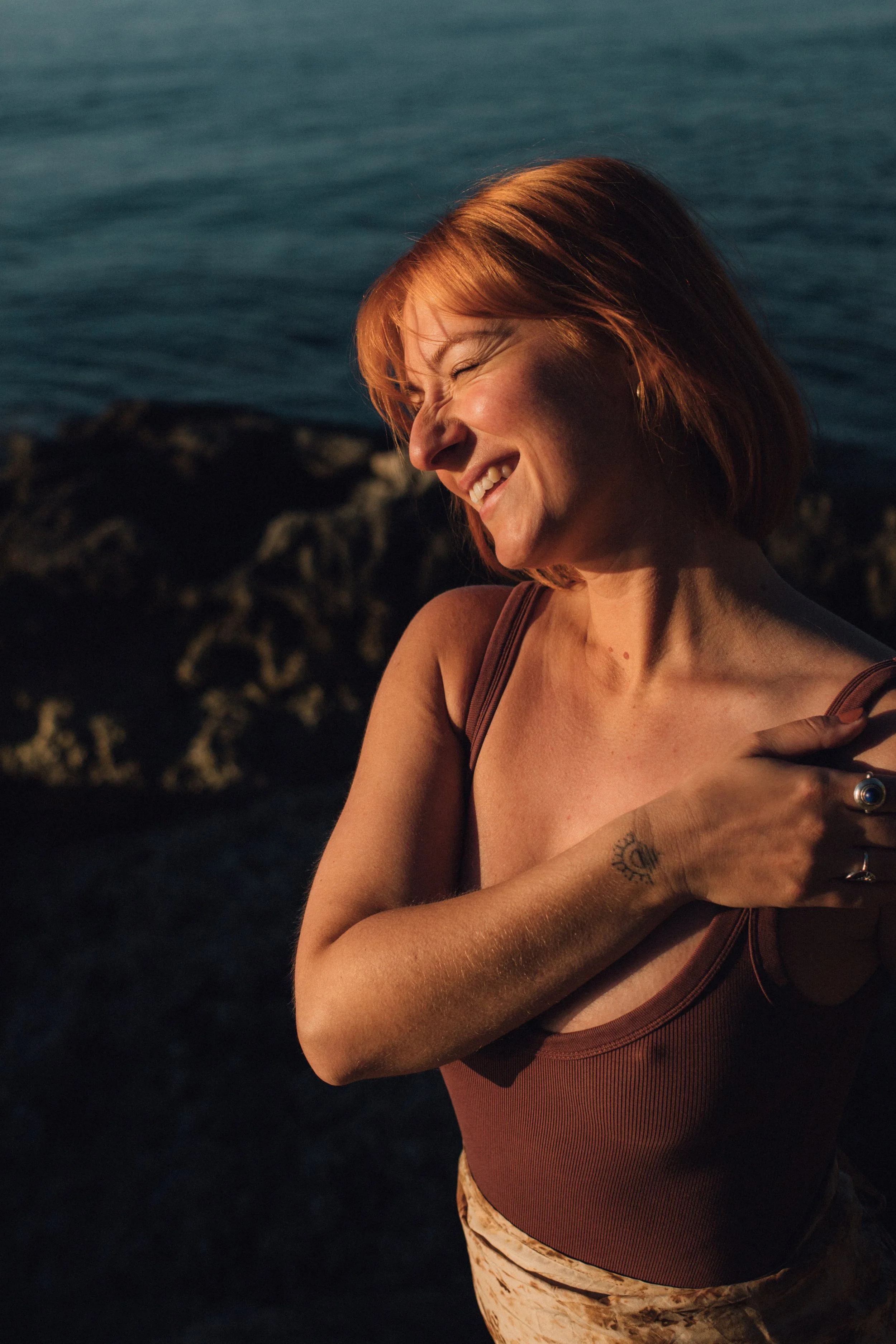 A woman with red hair smiling with her eyes closed, standing near water during sunset or sunrise, wearing a brown tank top and patterned pants, with a tattoo on her left arm.