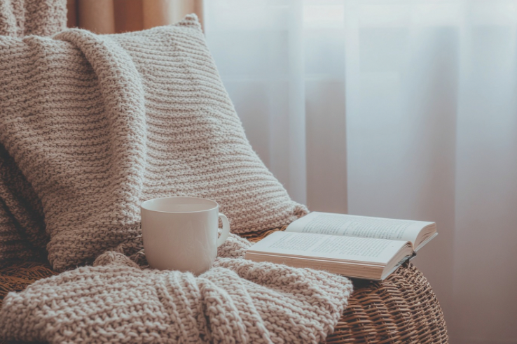 Cozy scene with a knitted blanket, pillows, a mug, and an open book on a wicker chair in front of a window with sheer curtains.