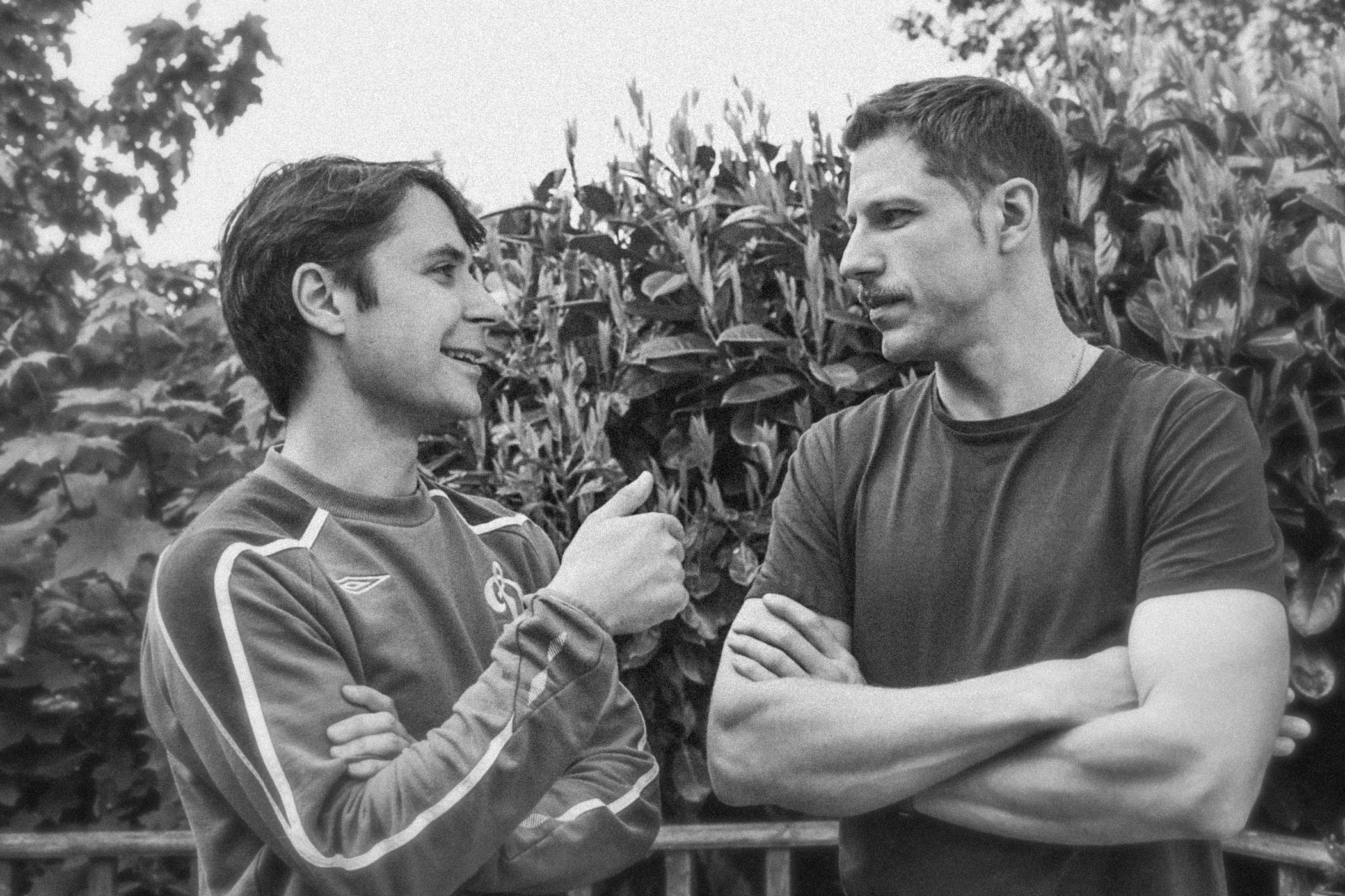 Two young men standing outdoors in front of a plant. One is smiling and holding a small object, while the other is looking at him with a serious expression. They are engaged in conversation.