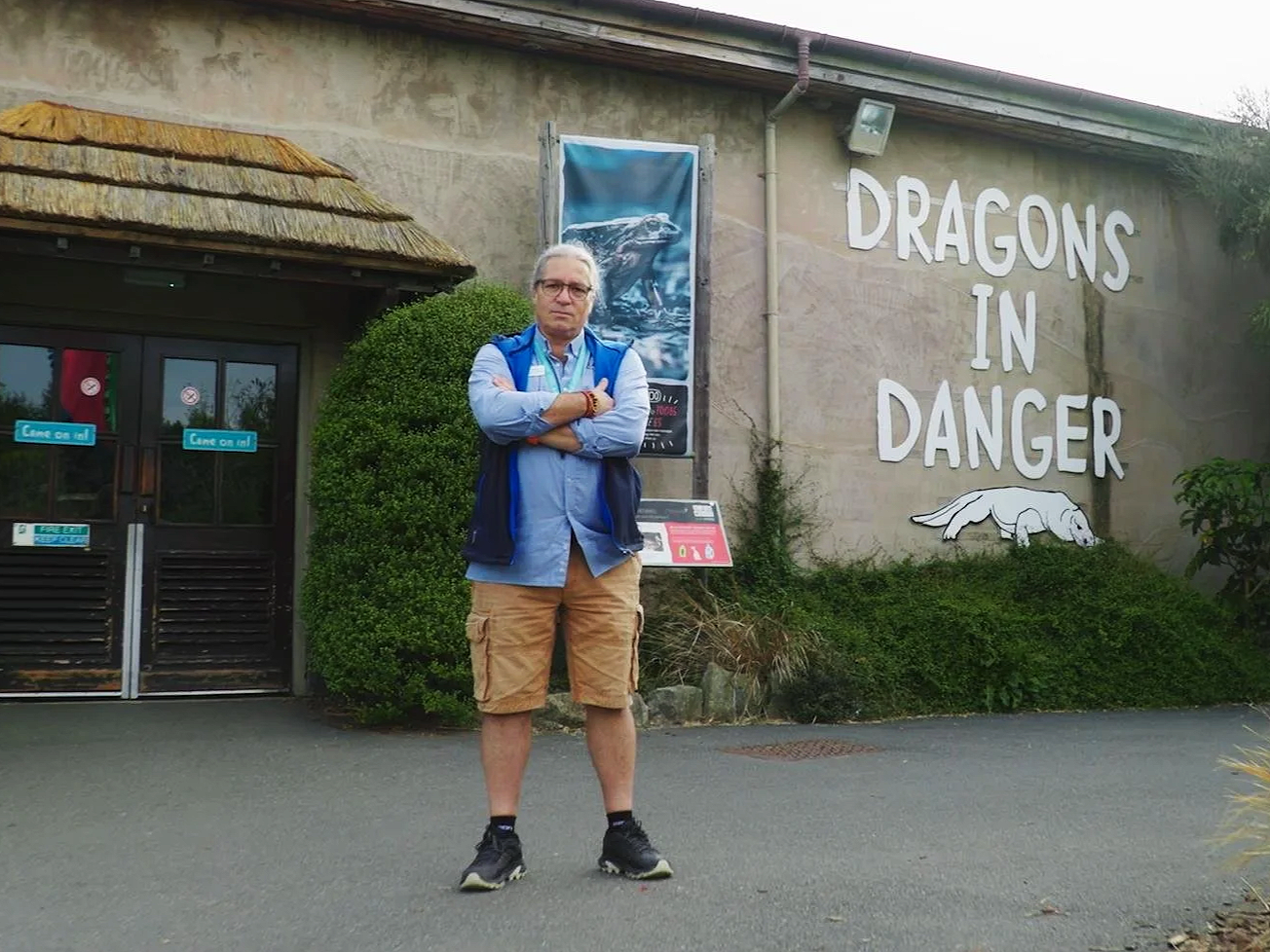 Man standing with arms crossed in front of a sign that reads "Dragons in Danger" at a zoo.