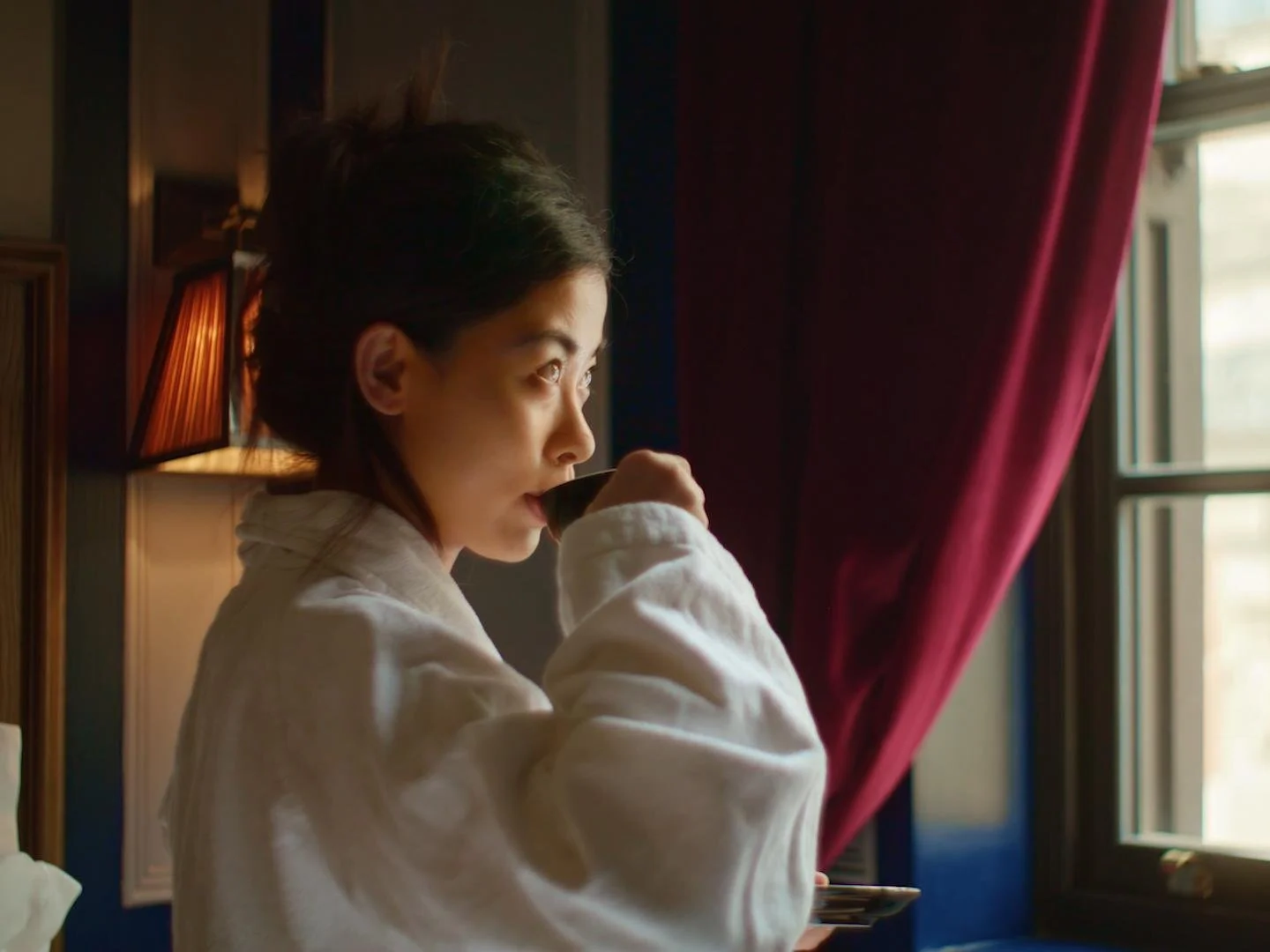 Young woman with short dark hair drinking from a cup while looking out of a window in a cozy room with red curtains.
