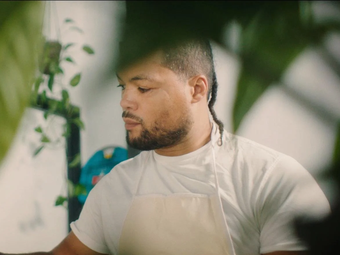 A man with short dark hair and a beard, wearing a white shirt and apron, painting in an art studio.