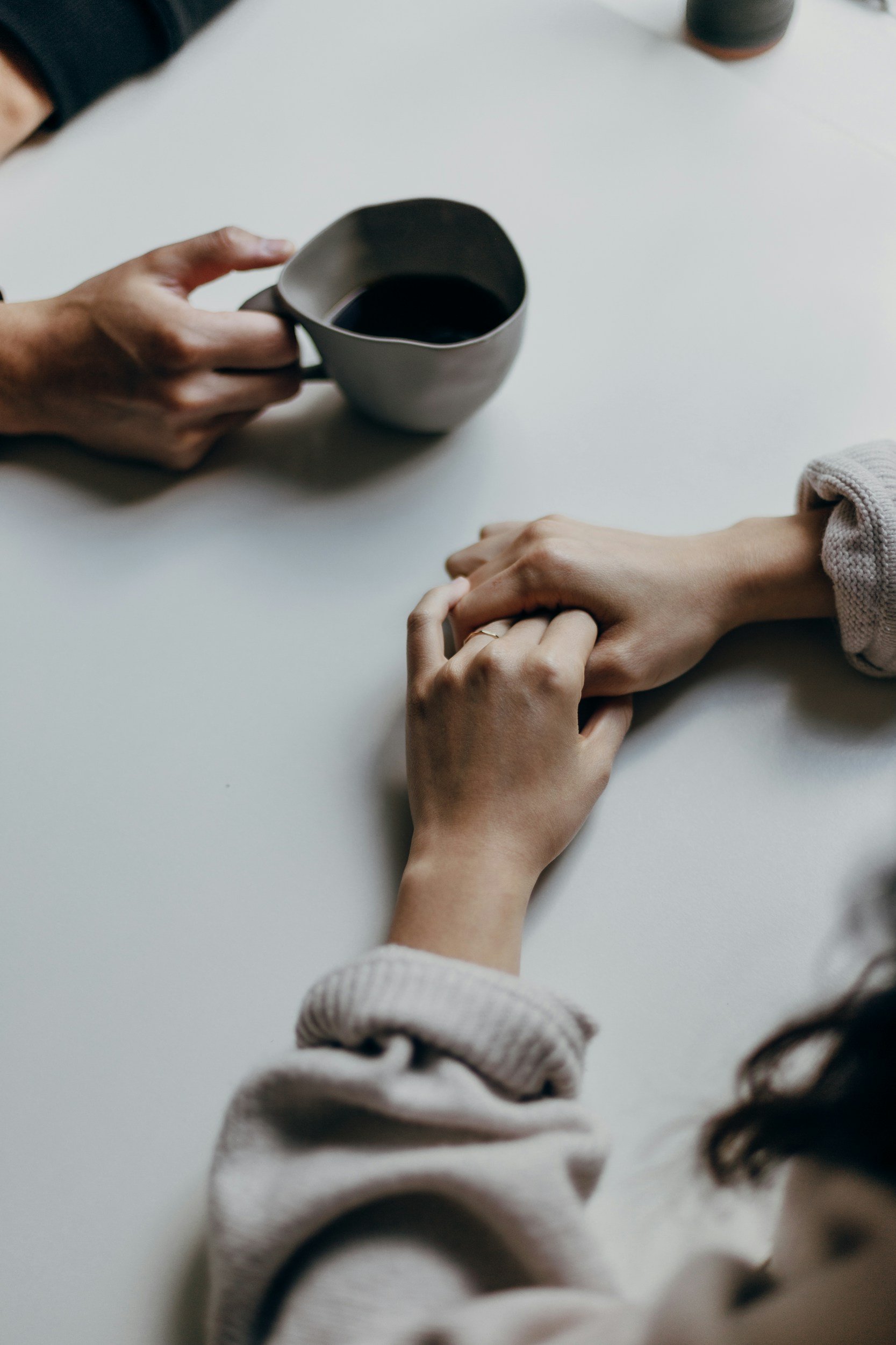 Two hands resting on a white table, one holding a mug filled with dark liquid, the other gently resting with fingers intertwined, with part of a person's arm visible wearing a light-colored sweater.