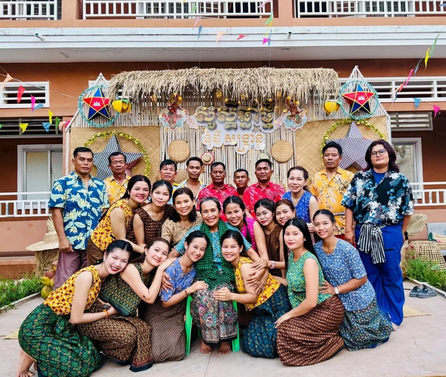 A group of people celebrating a cultural event in front of a decorated backdrop with traditional items, wearing colorful traditional clothing.