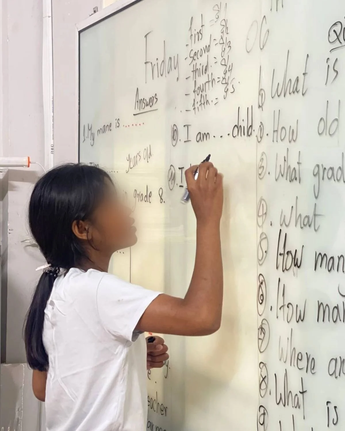 A young girl writing on a whiteboard with black marker, which displays answers to questions about age and grade, in a classroom setting.