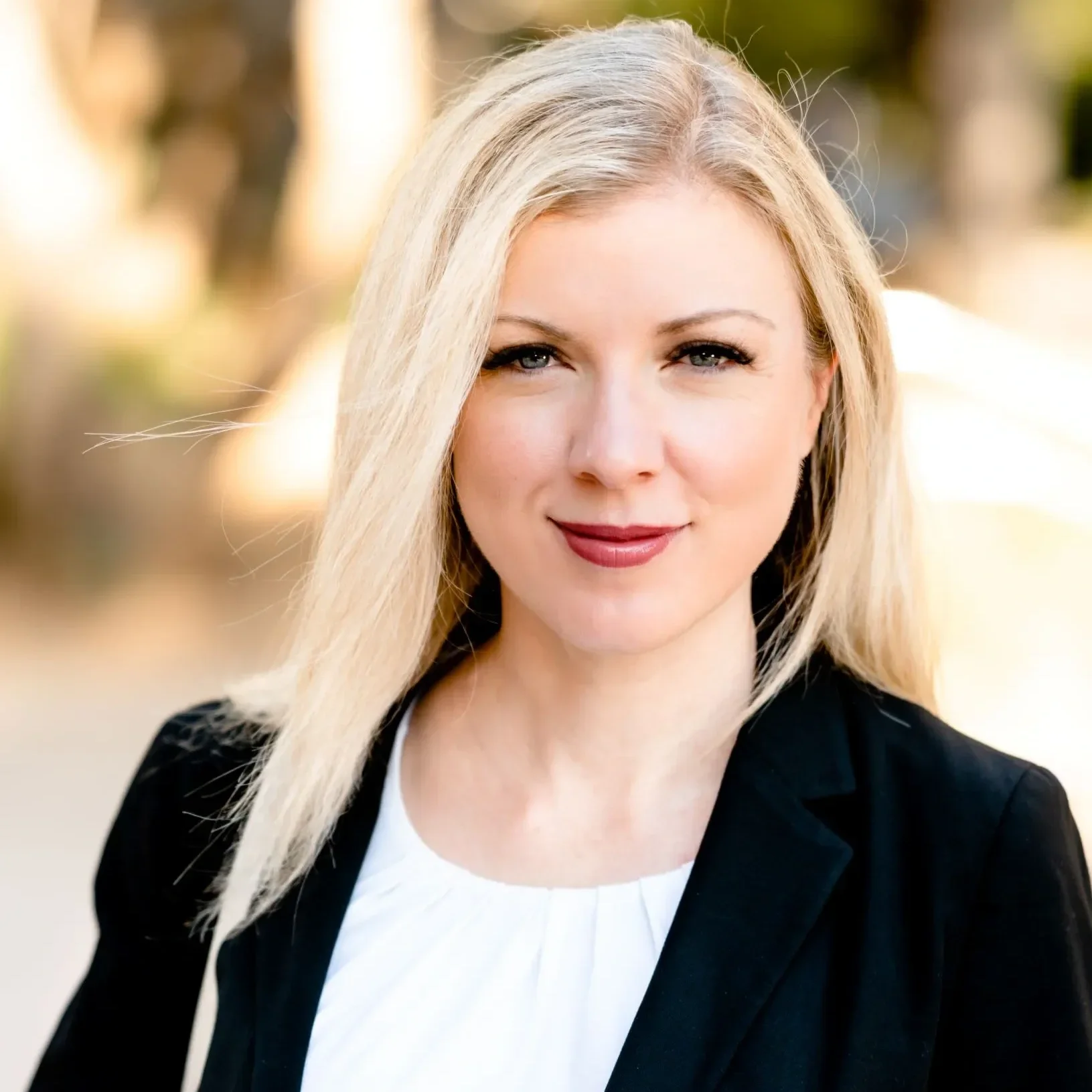 Close-up of a woman with blonde hair, blue eyes, and a slight smile, wearing a black blazer and white top, outdoors with blurred trees in the background.