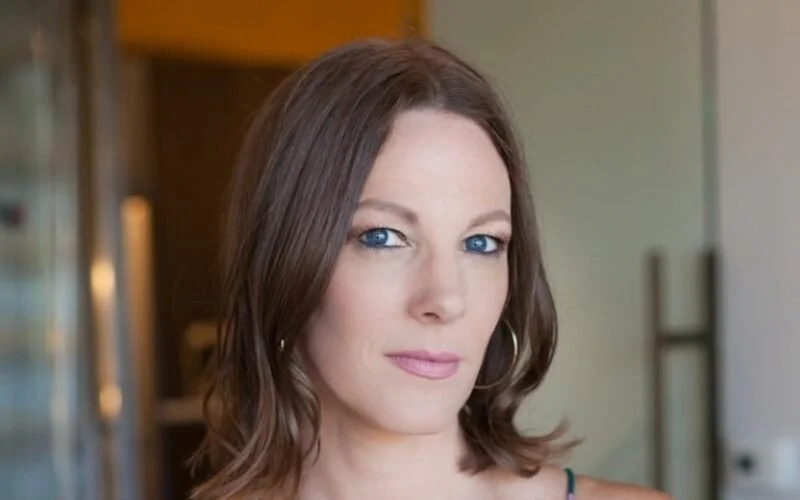Close-up of a woman with shoulder-length brown hair, blue eyes, and wearing hoop earrings, standing indoors with a blurred background.