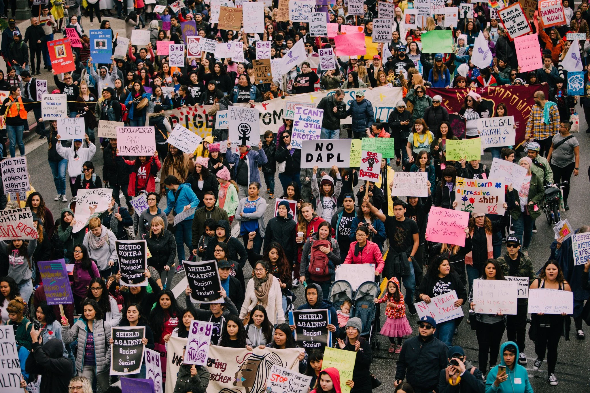 Large protest march with a diverse group of women and allies holding signs advocating for women's rights, LGBTQ+ rights, and social justice. Many wear pink, and signs contain messages about ending violence, discrimination, and supporting equality.