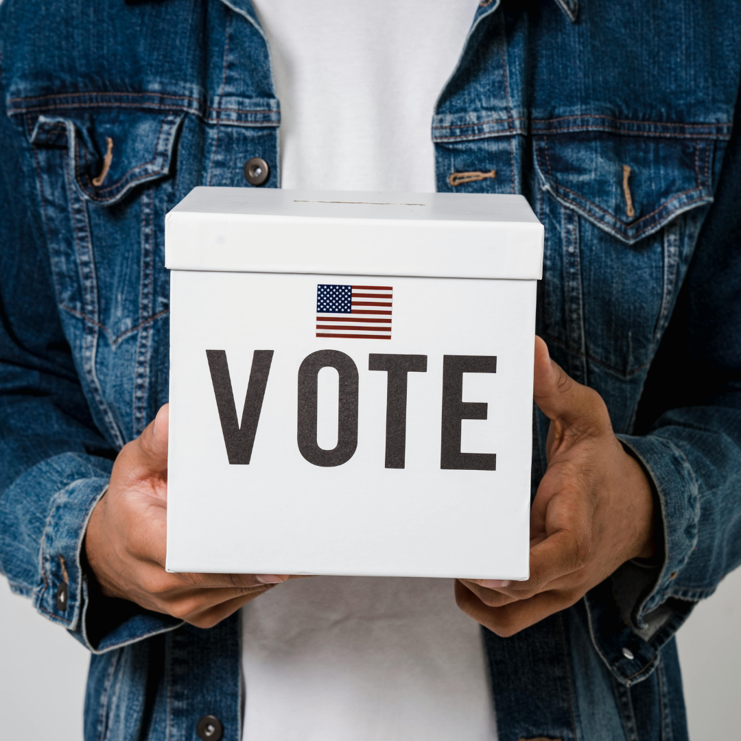 Person holding a white ballot box with 'VOTE' and an American flag on it, wearing a denim jacket.