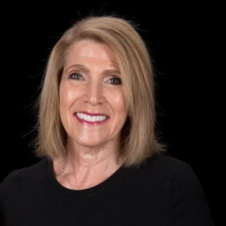 Headshot of a smiling middle-aged woman with shoulder-length blonde hair, wearing a black top against a black background.